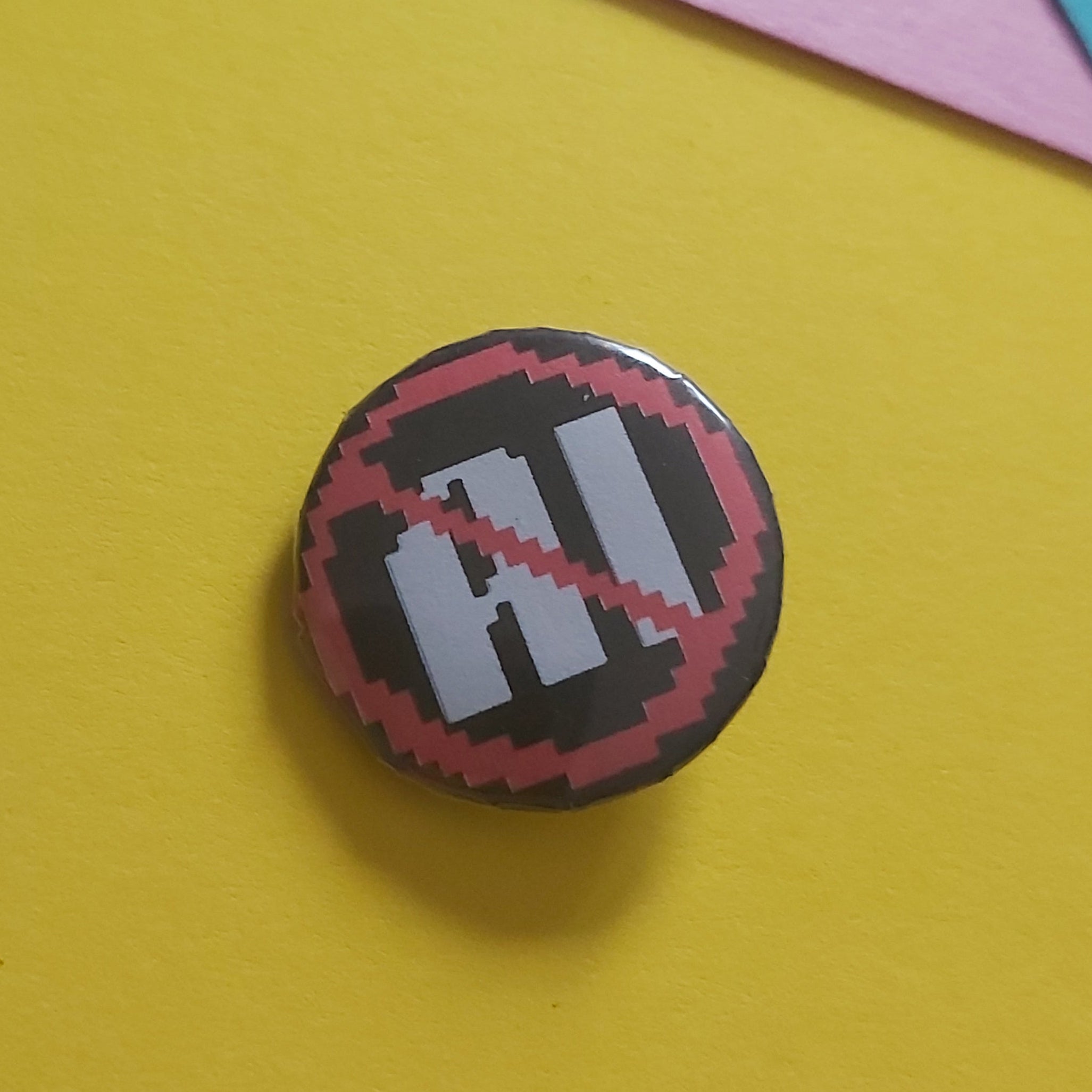 A small round pin badge with a black background, white stylised lettering, and a red diagonal slash across it, placed on a bright yellow surface with pink and blue paper accents in the background.