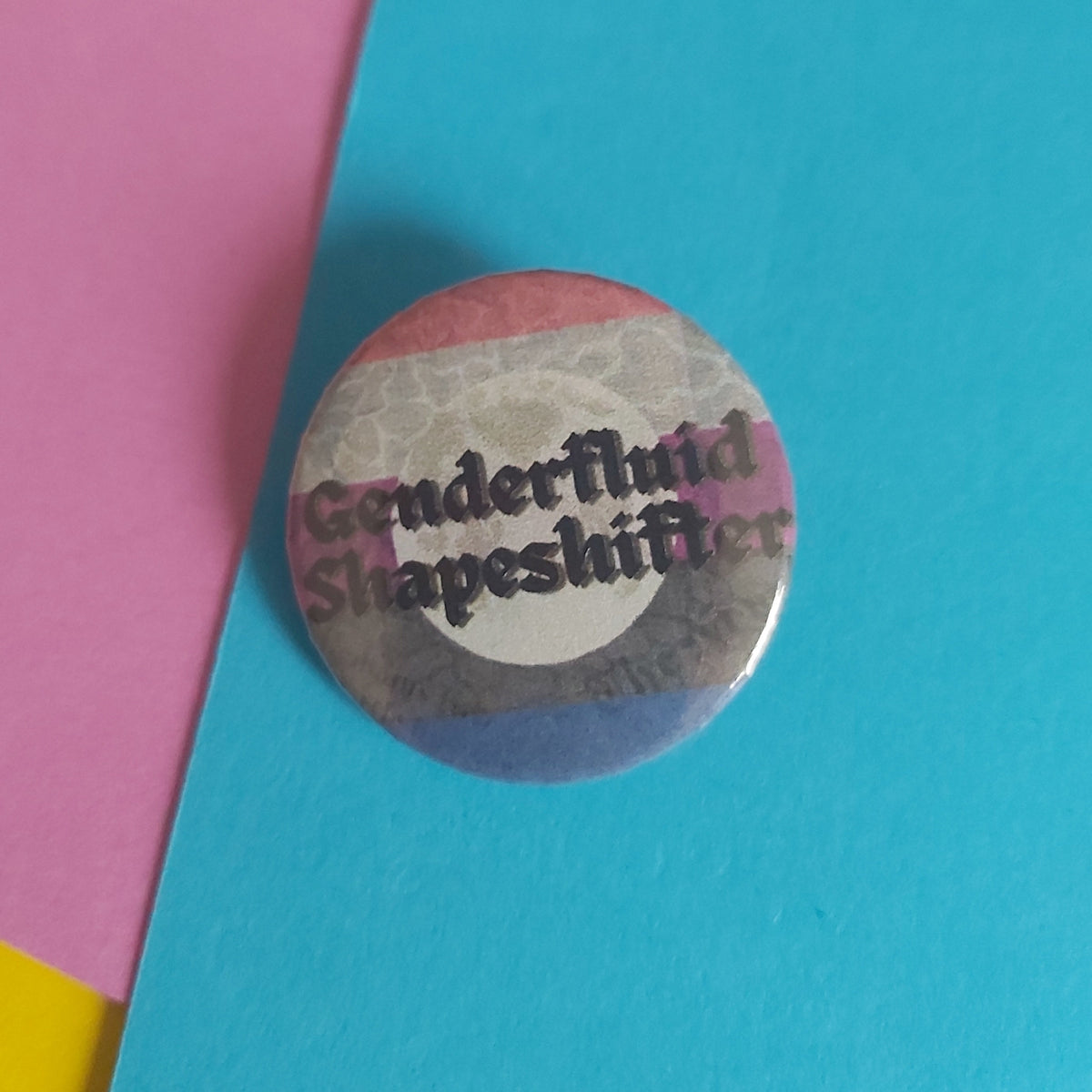 A round pin-back button badge featuring the text 'Genderfluid Shapeshifter' in black script over a distressed pink, white, gray, and blue flag design, placed on a background of pink, blue, and yellow paper.