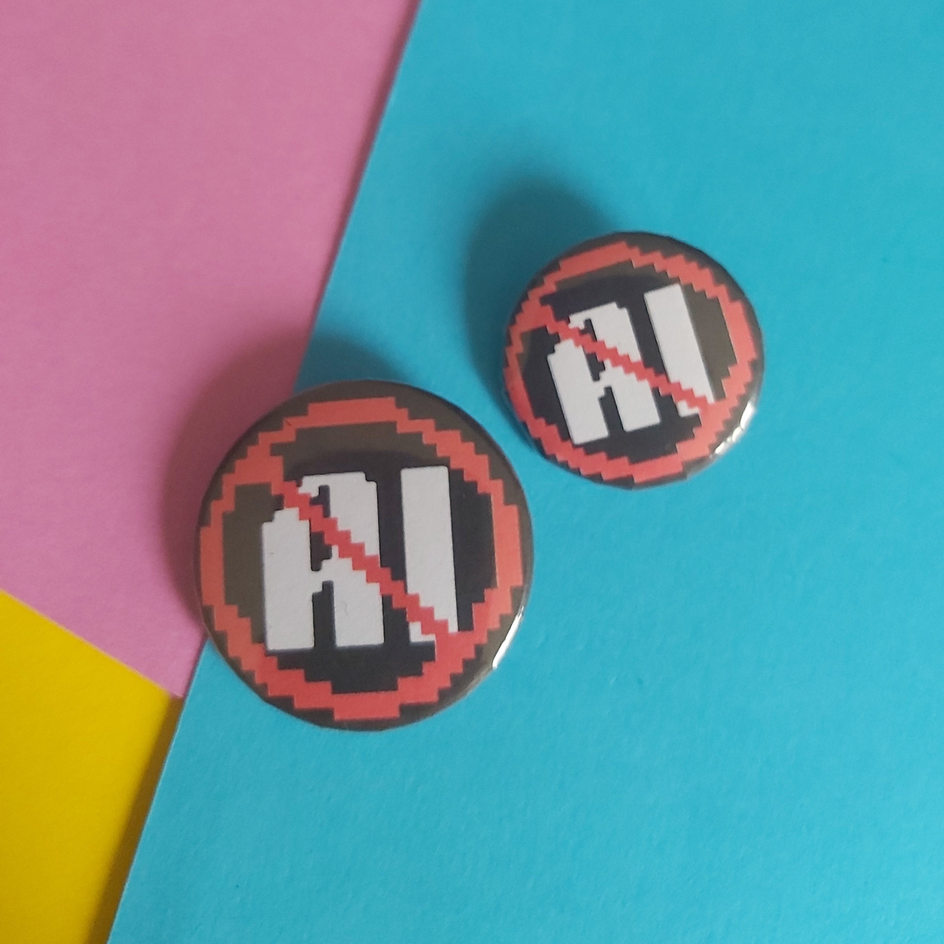 Two round button badges with a 'no AI' pixel-art design, displayed on a diagonally split pink, blue, and yellow background.