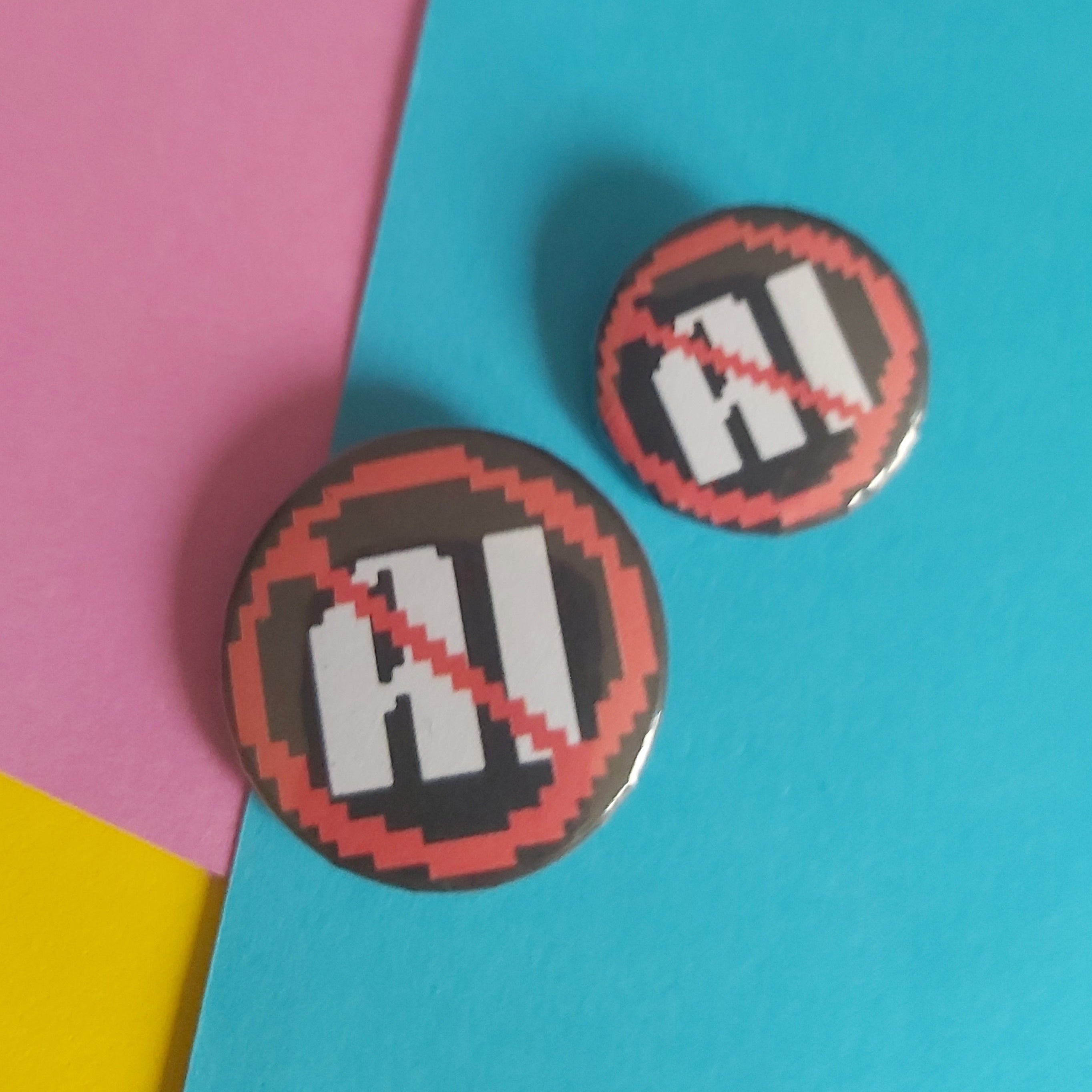 Two round pin-back buttons featuring a pixelated 'AI' crossed out with a red diagonal slash, displayed on a diagonally split pink, blue, and yellow background.