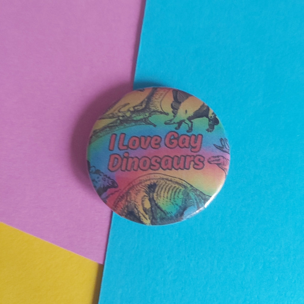 A round pin-back button badge featuring a colourful dinosaur illustration and the text 'I Love Gay Dinosaurs' in red lettering, placed on a diagonally split pink, yellow, and blue paper background.