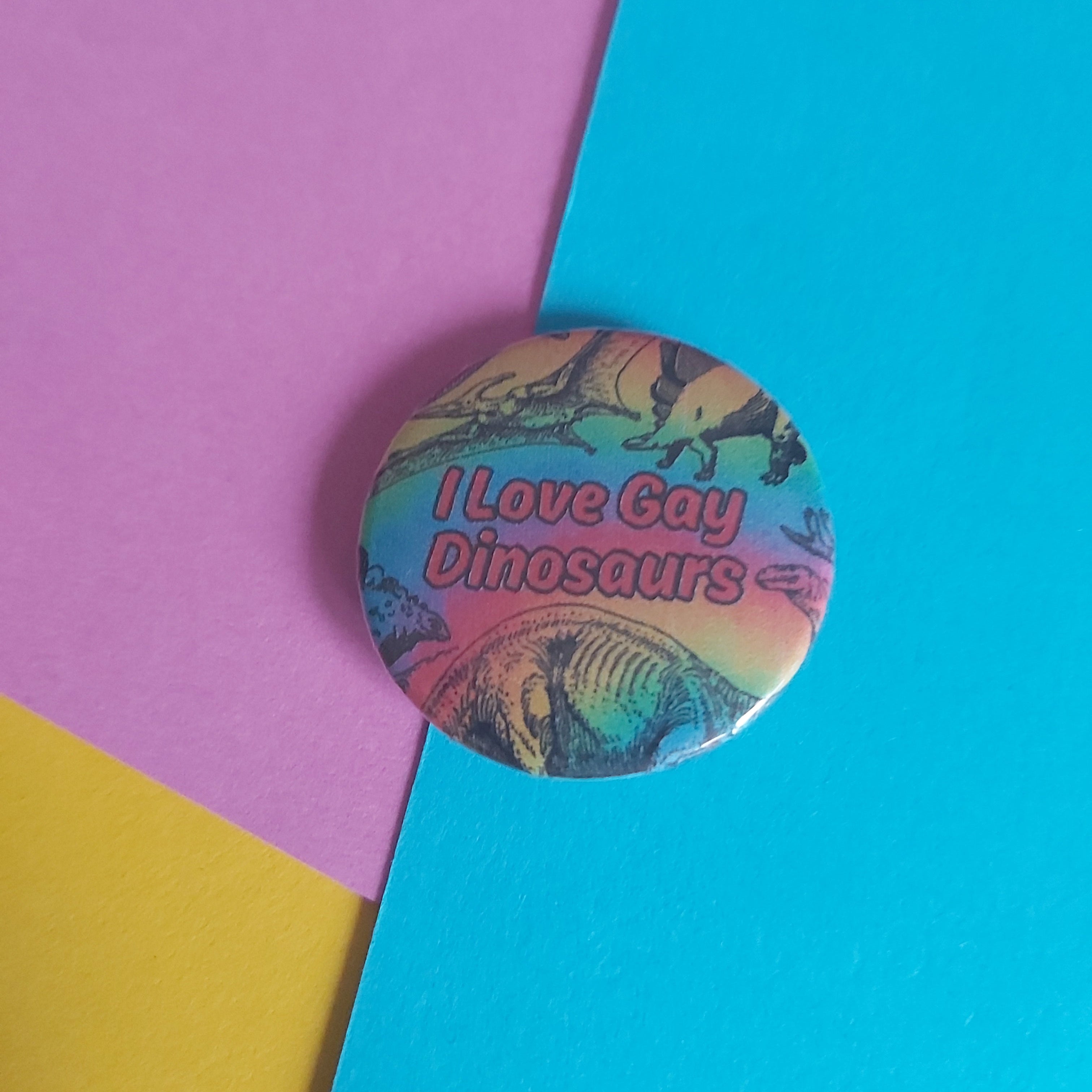 A round pin-back button badge featuring the text 'I Love Gay Dinosaurs' in red lettering over a colourful rainbow dinosaur illustration, placed on a diagonally split background of purple, yellow, and bright blue paper.