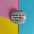 A round pinback button with a grey and light blue striped demiboy pride flag design and the text 'SOMETIMES A BOY. SOMETIMES NO GENDER.