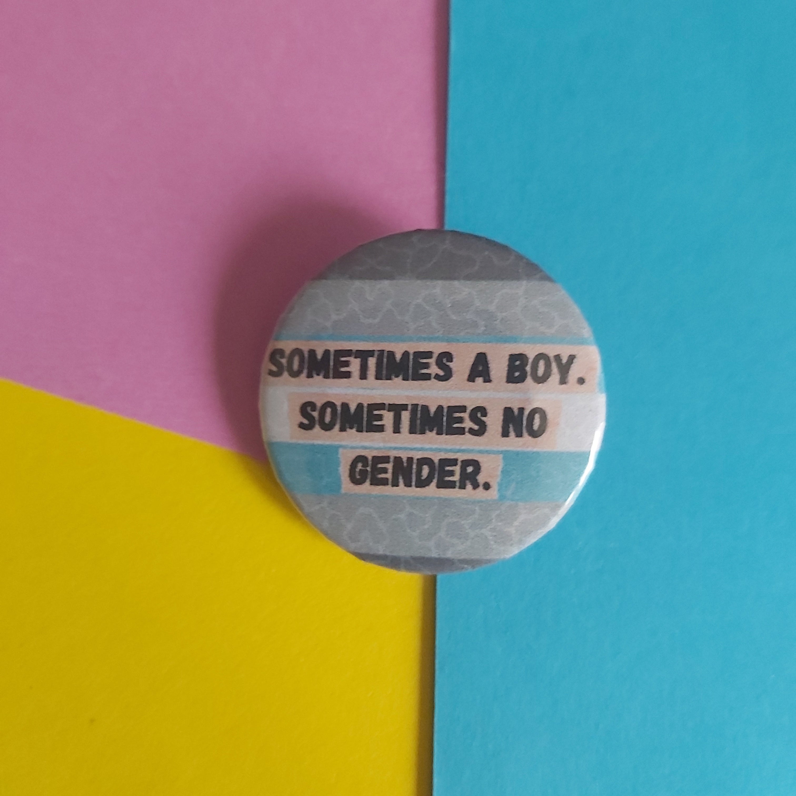 A round pinback button with a grey and light blue striped demiboy pride flag design and the text 'SOMETIMES A BOY. SOMETIMES NO GENDER.