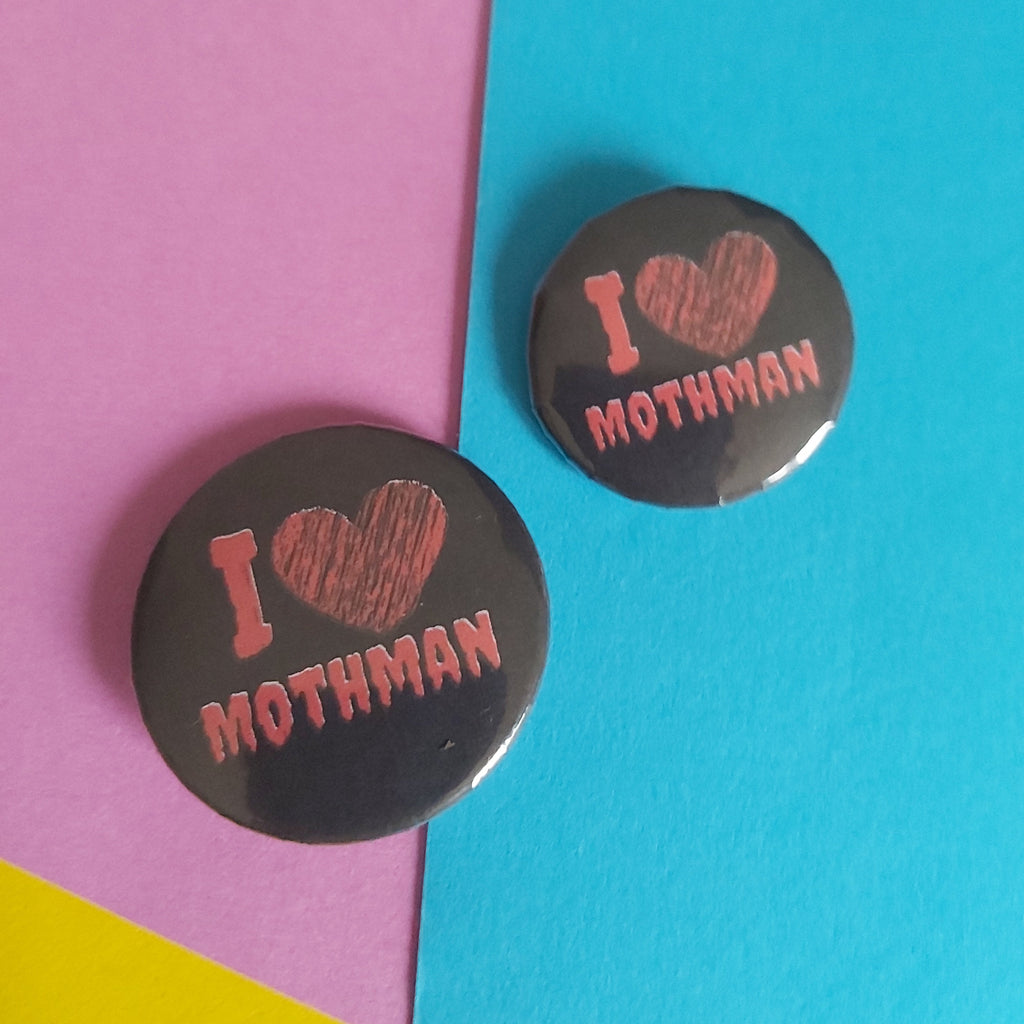 Two round black pin-back buttons with a pink 'I [heart] MOTHMAN' design, displayed on a split pink and blue background with a small yellow corner.