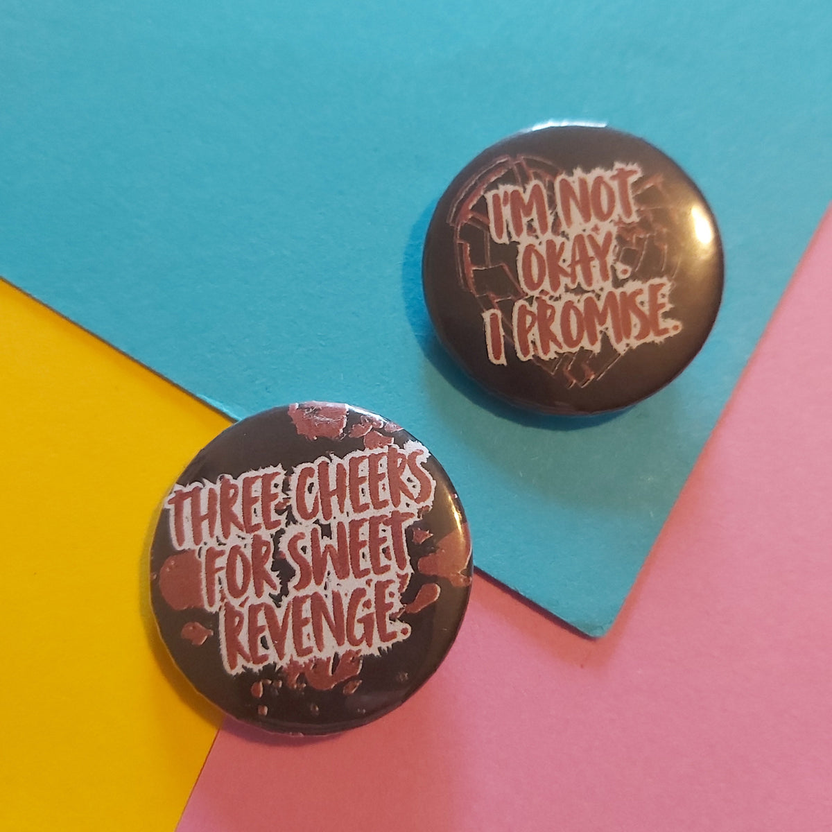Two round pin-back buttons displayed on a colourful geometric background. One button is black with red splatter graphics and white text reading 'THREE CHEERS FOR SWEET REVENGE'.