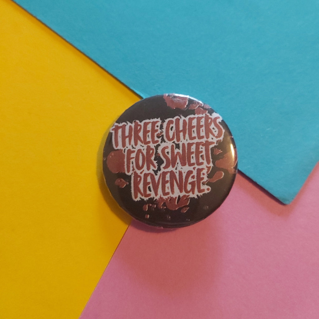 A round pin badge with the text 'THREE CHEERS FOR SWEET REVENGE' in white and red lettering, set against a dark background with red splatter details, placed on a diagonally split yellow, blue, and pink paper background.