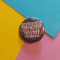 A round pin badge with the text 'THREE CHEERS FOR SWEET REVENGE' in white and red lettering, set against a dark background with red splatter details, placed on a diagonally split yellow, blue, and pink paper background.