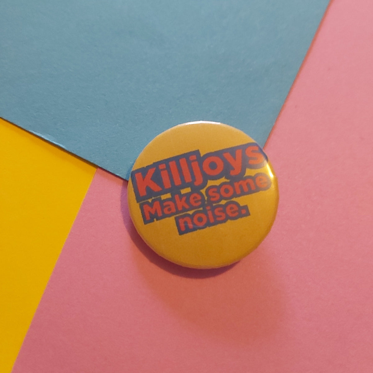 A round yellow pinback button with the text 'Killjoys Make some noise.' in red and blue, placed on a colorful background of pink, blue, and yellow paper.