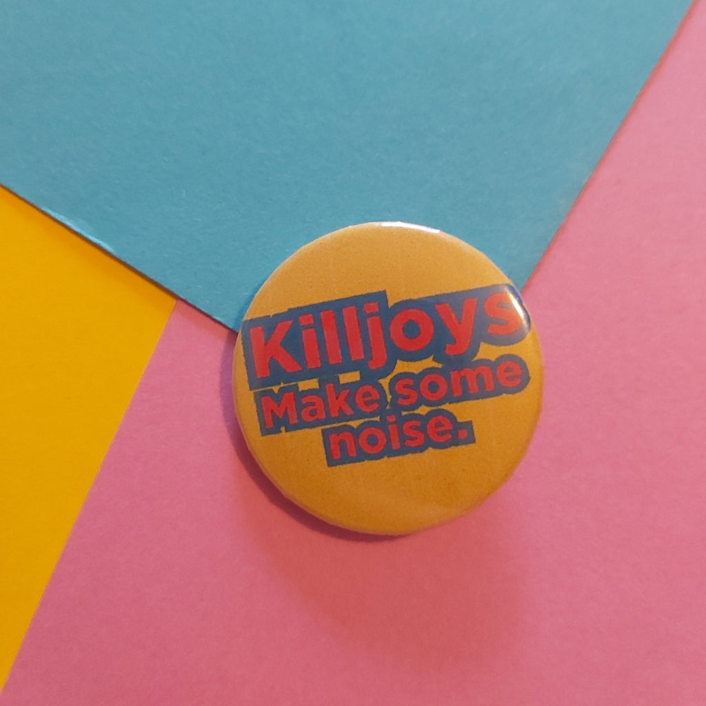A round pin badge with the text 'Killjoys Make some noise.' in red and blue lettering on a yellow background, placed on a colorful geometric paper backdrop of pink, blue, and yellow.