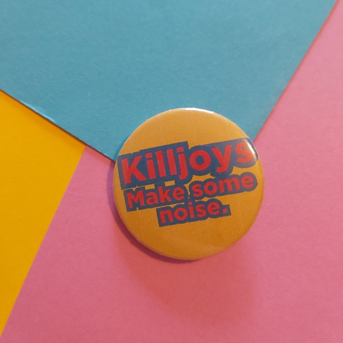 A round pin badge with the text 'Killjoys Make some noise.' in red and blue lettering on a yellow background, placed on a colorful geometric paper backdrop of pink, blue, and yellow.