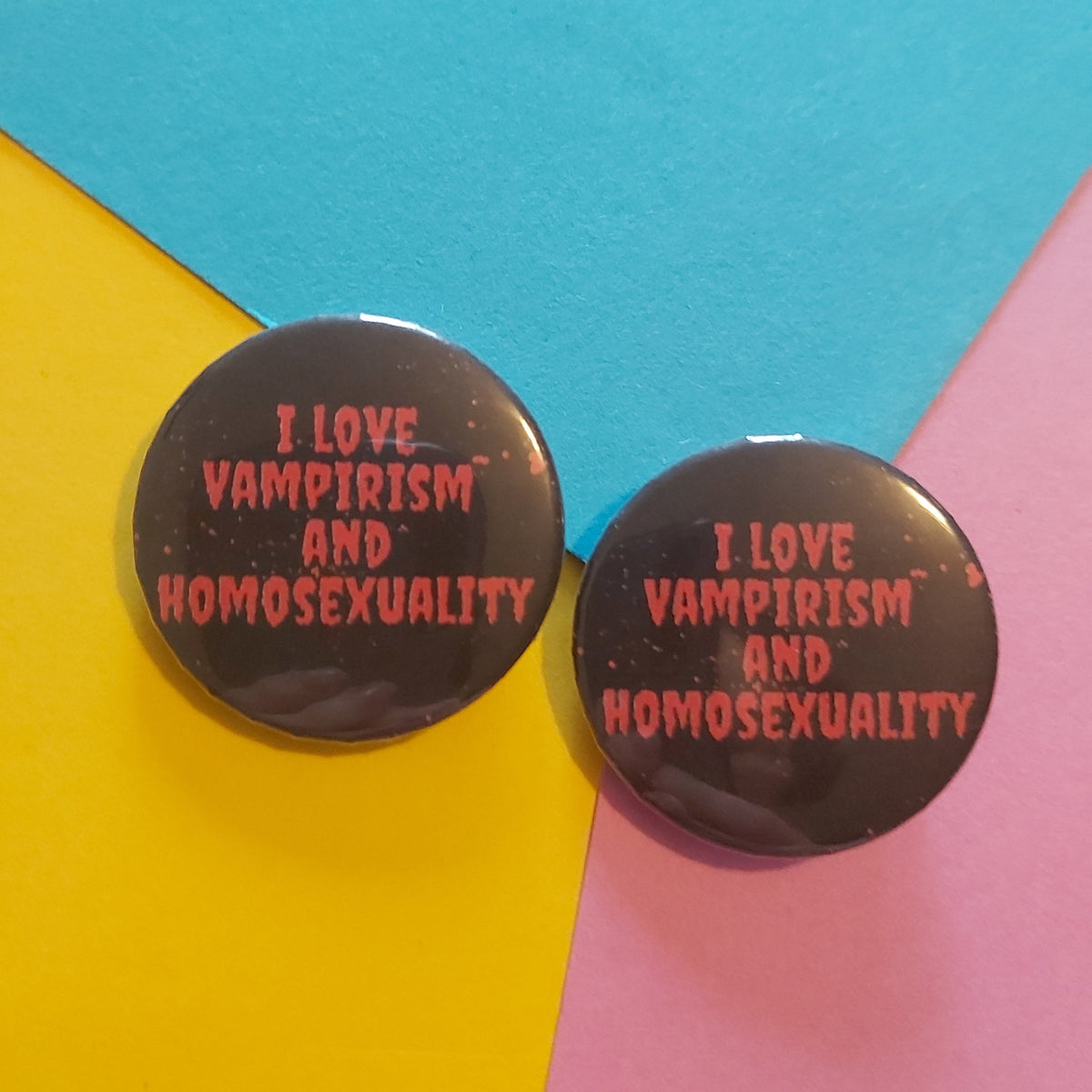 Two round black pin-back buttons with red text reading 'I LOVE VAMPIRISM AND HOMOSEXUALITY' displayed on a colourful background of yellow, blue, and pink paper.