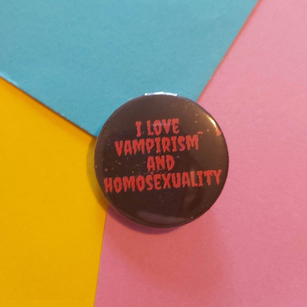 A round pinback button with red text reading 'I LOVE VAMPIRISM AND HOMOSEXUALITY' on a glossy dark background, placed on a colorful geometric paper backdrop with blue, yellow, and pink sections.