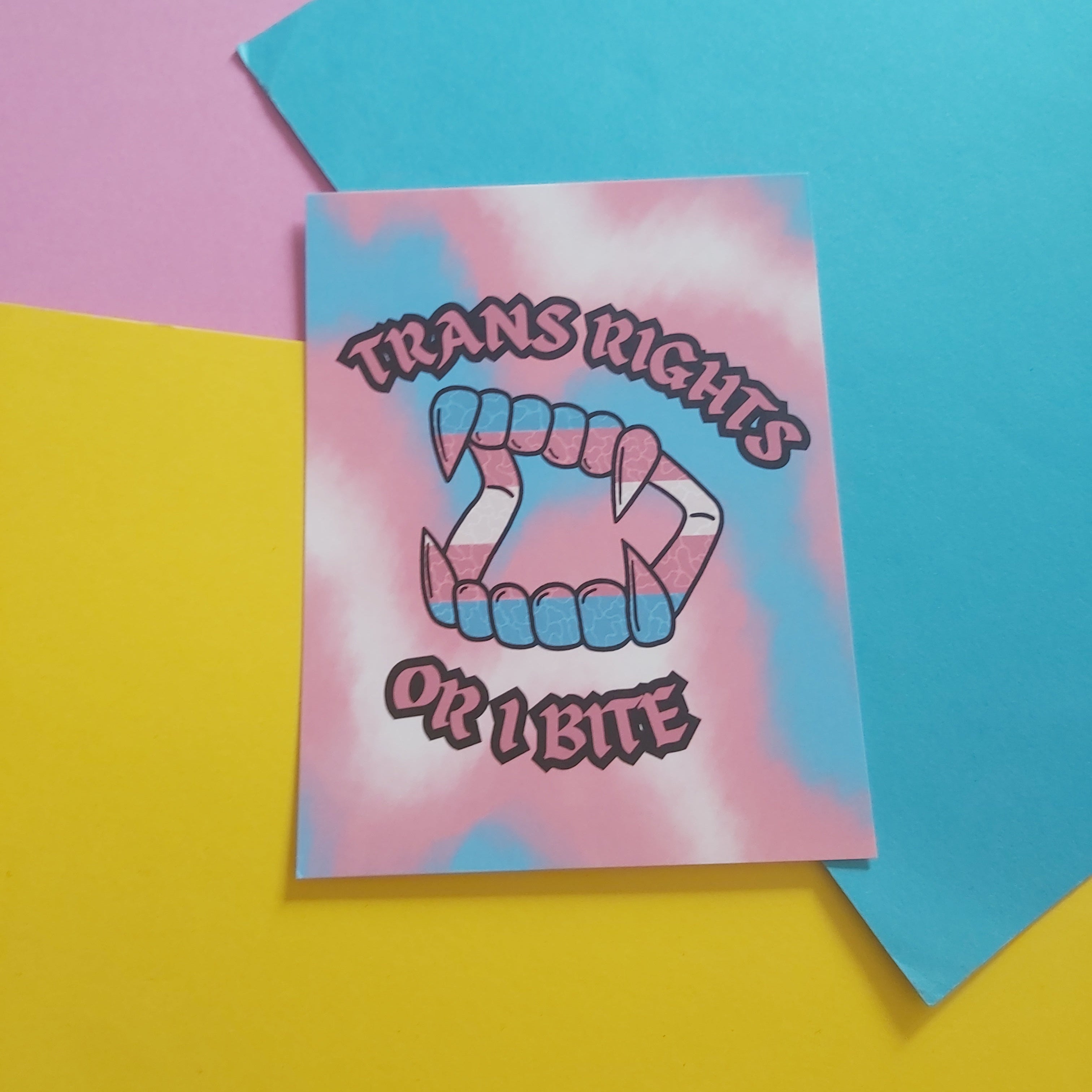A print with a pink, white, and blue tie-dye background, featuring a fanged mouth symbol and the text 'TRANS RIGHTS OR I BITE' in stylised lettering. The card is photographed on a flat lay of pastel pink, yellow, and blue paper.