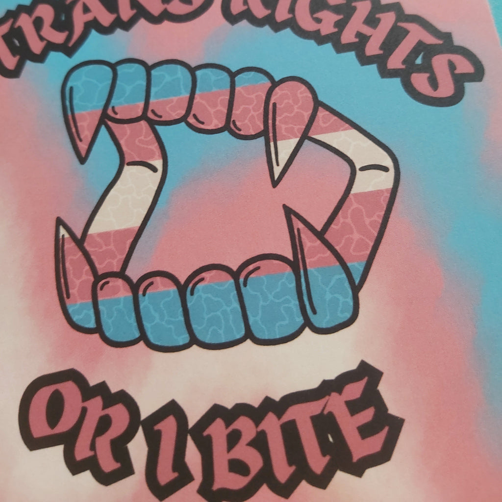 A close-up of a graphic print featuring stylised fangs outlined in black, filled with the pink, white, and blue stripes of the transgender flag. The text reads 'TRANS RIGHTS OR I BITE' at the bottom, set against a pink and blue tie-dye background.