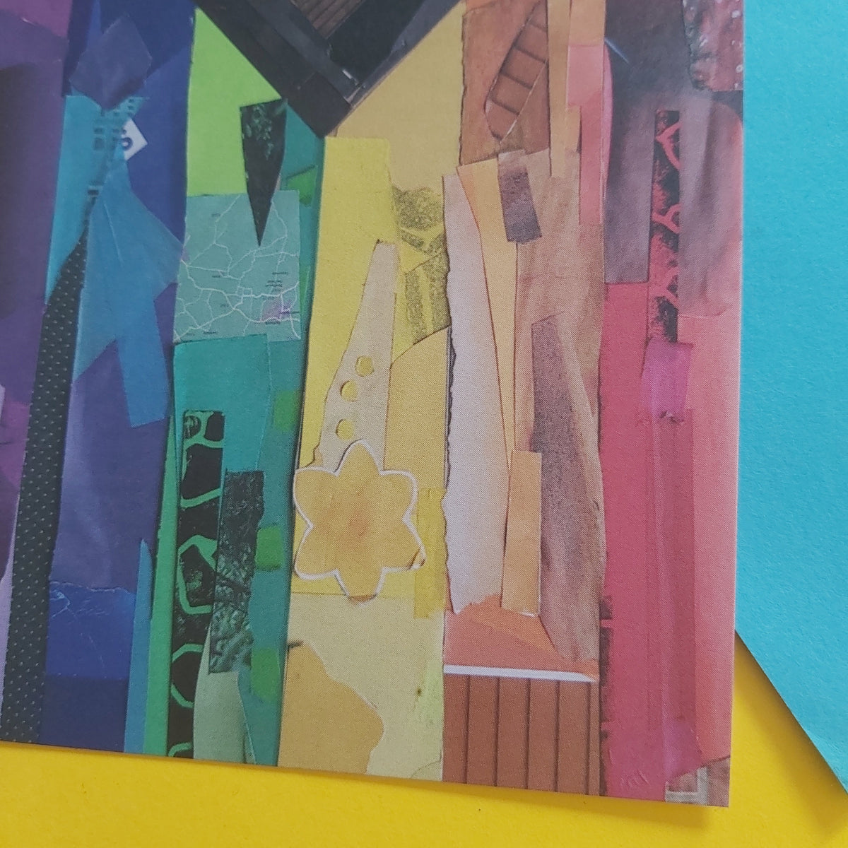 A close-up of a colourful collage artwork made from vertical strips of paper in blue, green, yellow, beige, brown, and pink, placed on a bright yellow surface with a blue paper sheet visible at the right edge.