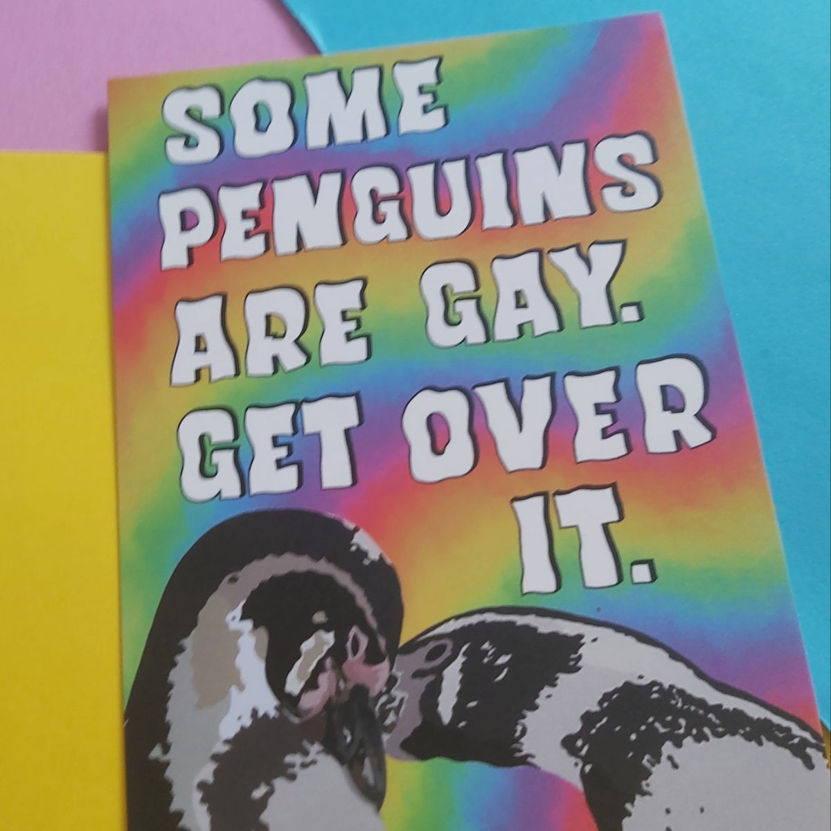 A rectangular print with a rainbow tie-dye background, large white text reading “SOME PENGUINS ARE GAY. GET OVER IT.