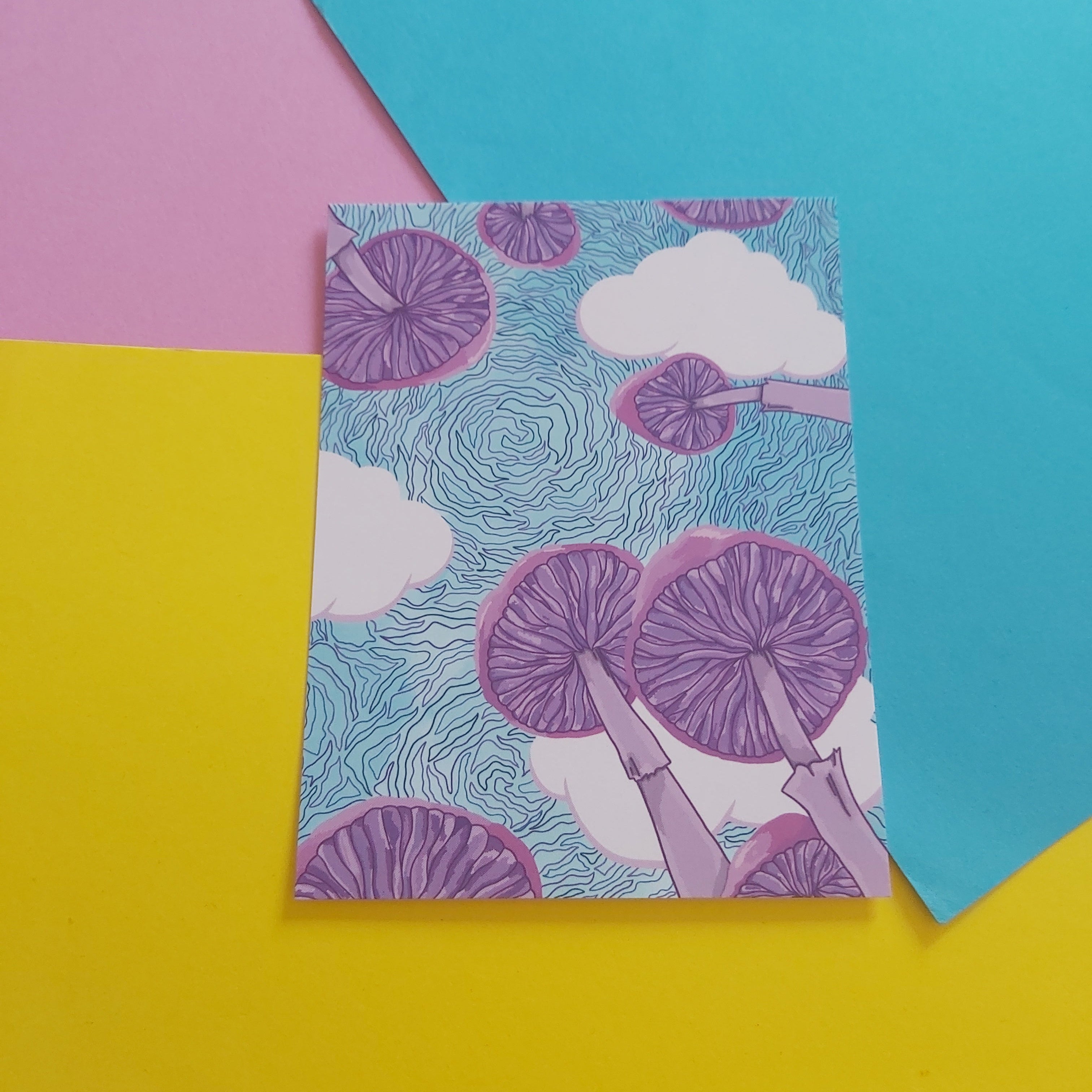 A rectangular art print featuring a whimsical illustration of purple mushrooms and white clouds on a light blue background, styled on a colourful flat-lay backdrop of pink, yellow, and blue paper.