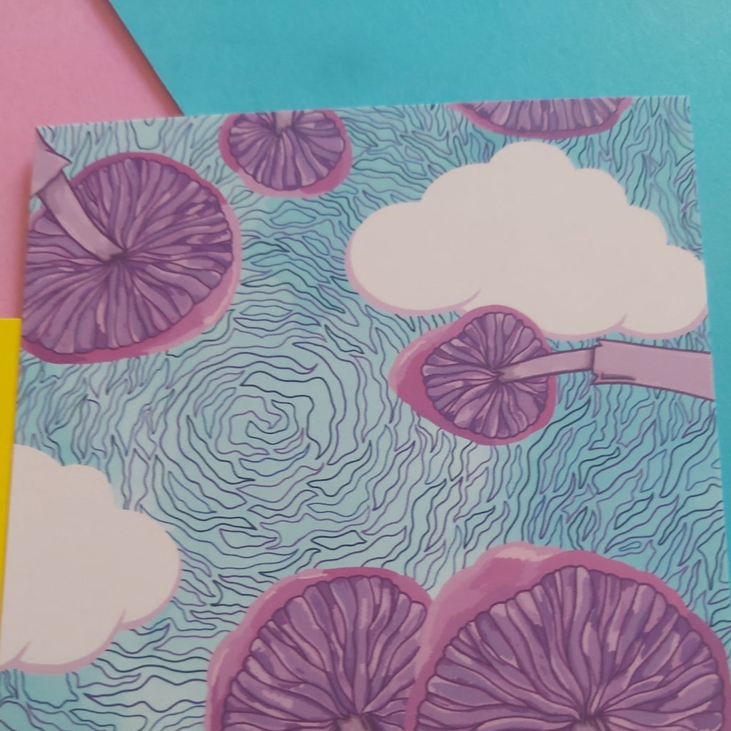 A close-up of a colourful printed surface featuring purple mushroom illustrations, white clouds, and a swirling light blue pattern, with pink, yellow, and teal paper edges visible around it.