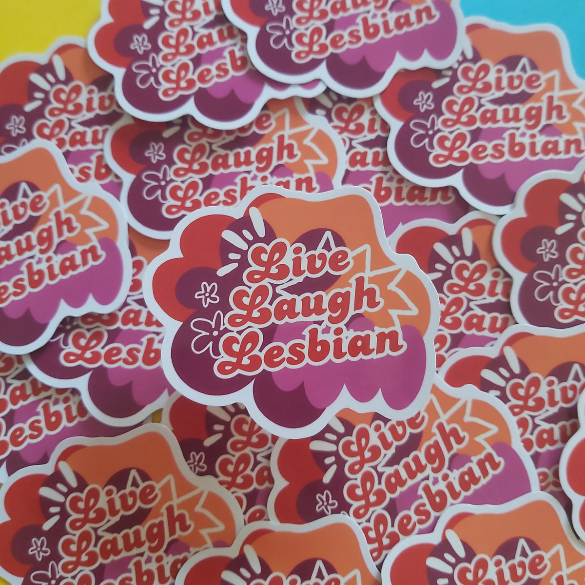 A pile of die-cut stickers featuring the phrase 'Live Laugh Lesbian' in stylised pink and orange lettering over a retro floral background, arranged on a bright yellow and blue surface.