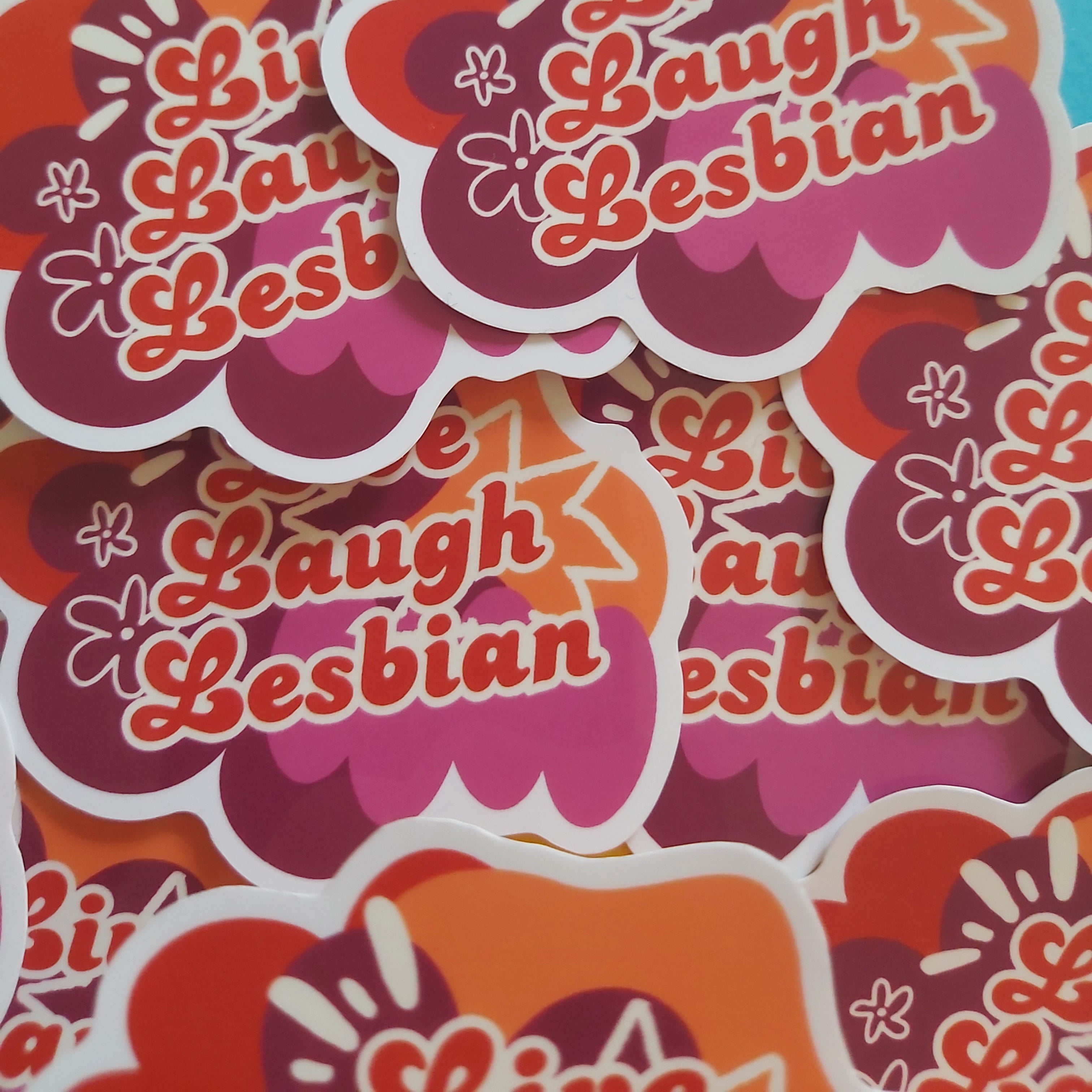 A pile of die-cut stickers featuring retro-style pink, orange, and red shapes with the text 'Live Laugh Lesbian' in red script, photographed on a colorful pastel background.