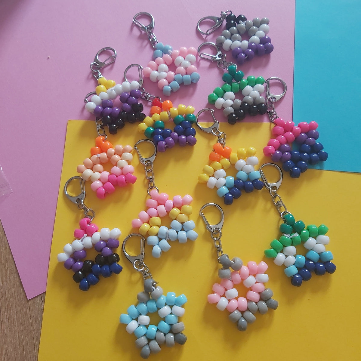 A group of handmade beaded keychains arranged on pink, yellow, and blue paper sheets. The keychains feature various colorful bead patterns including rainbow gradients, stripes, and geometric shapes, attached to silver lobster clasps.