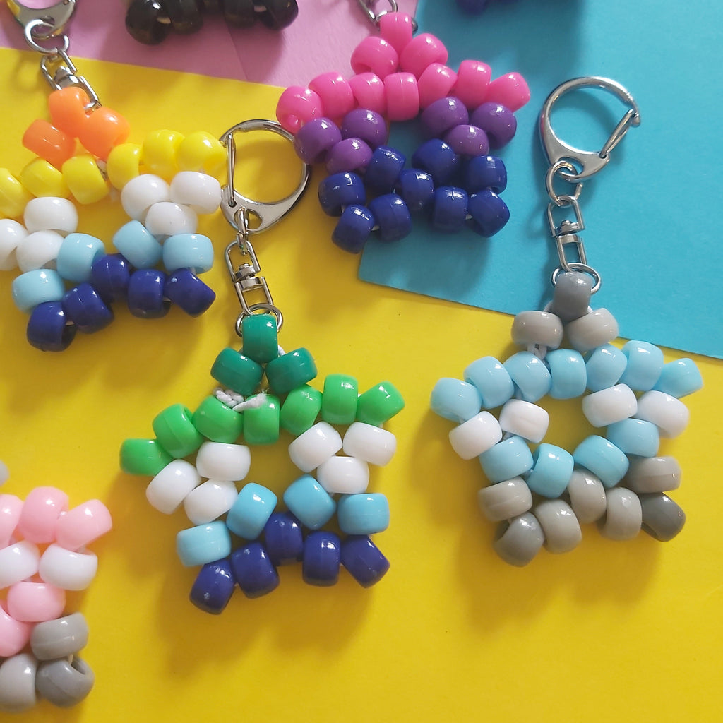 Several handmade beaded snowflake keychains in various color combinations are arranged on a bright yellow and blue background.