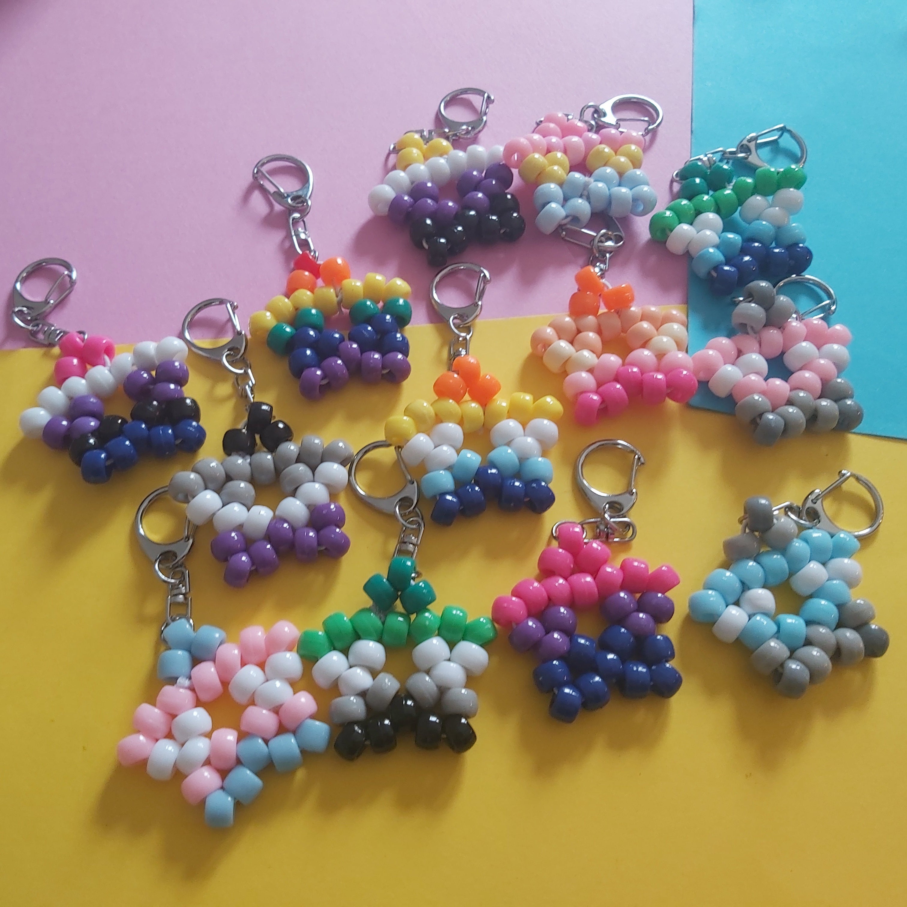 A group of handmade beaded keychains arranged on pink, yellow, and blue paper backgrounds. The keychains feature various colorful bead patterns including rainbow, striped, and geometric designs in pink, blue, purple, green, yellow, white, and black.