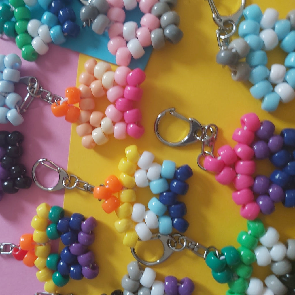 Several beaded keychains with silver lobster clasp closures arranged on a split pink and yellow background. The keychains feature colorful bead patterns including rainbow stripes, floral motifs, and geometric shapes in pink, blue, yellow, green, purple, white, and black.
