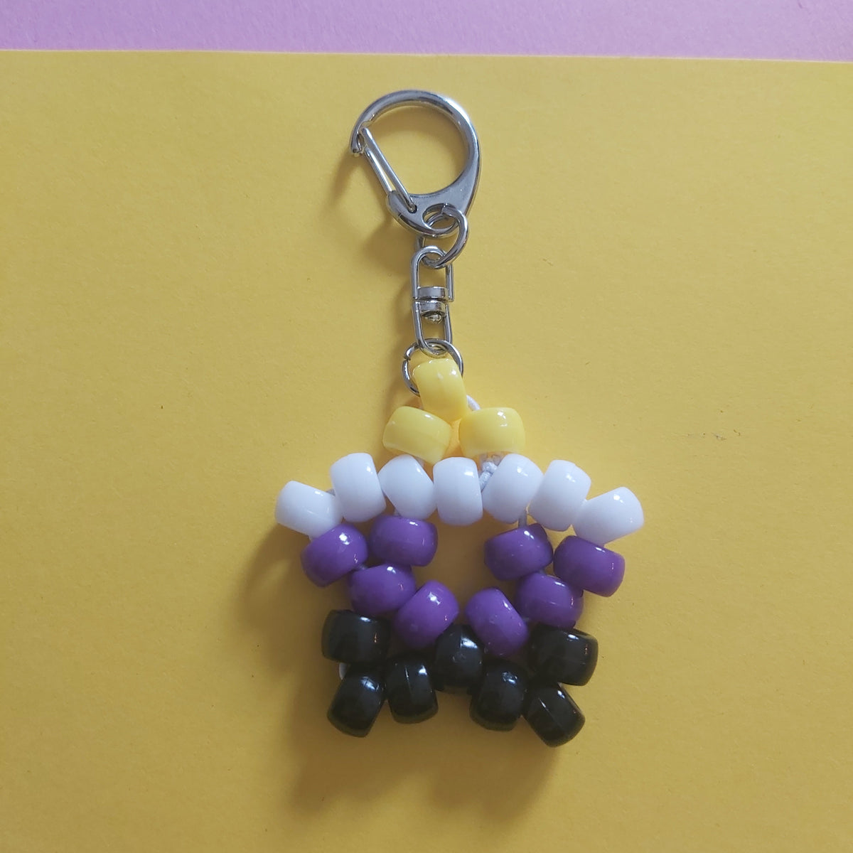 A beaded keychain charm shaped like a star, made with white, purple, and black beads representing the non-binary flag, attached to a silver metal lobster clasp, set against a split yellow and purple background.