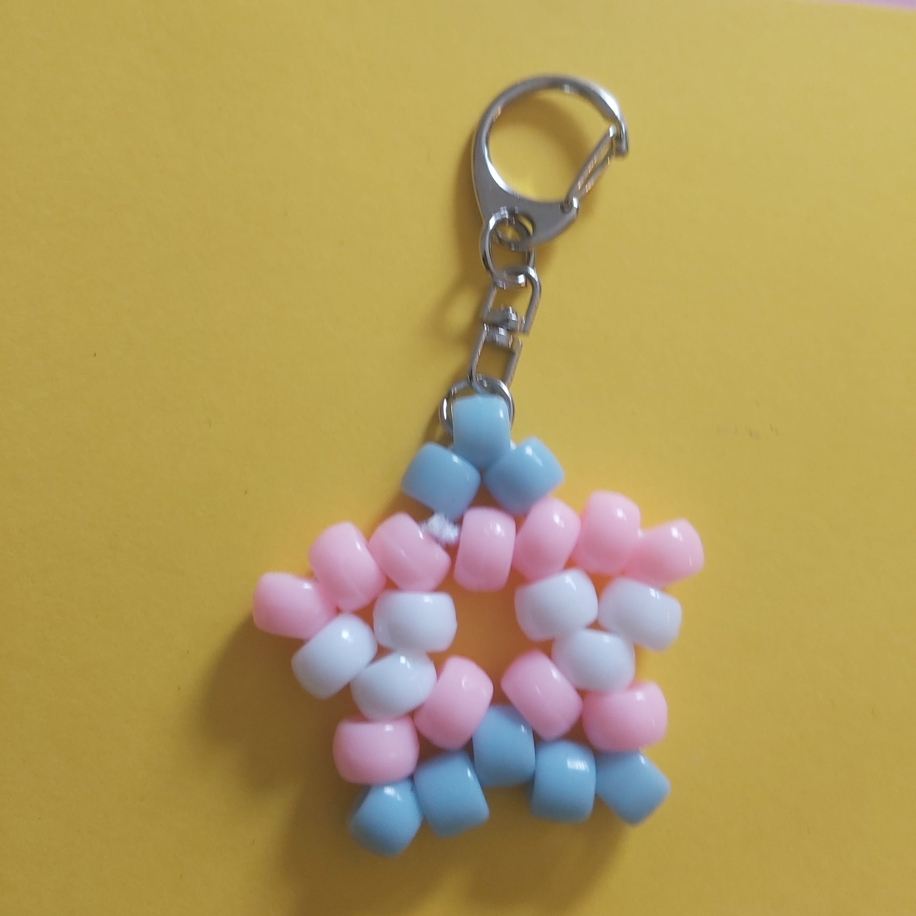 A handmade beaded keychain charm shaped like a star, featuring pink, light blue, and white beads attached to a silver metal lobster clasp, set against a split yellow and pink background.