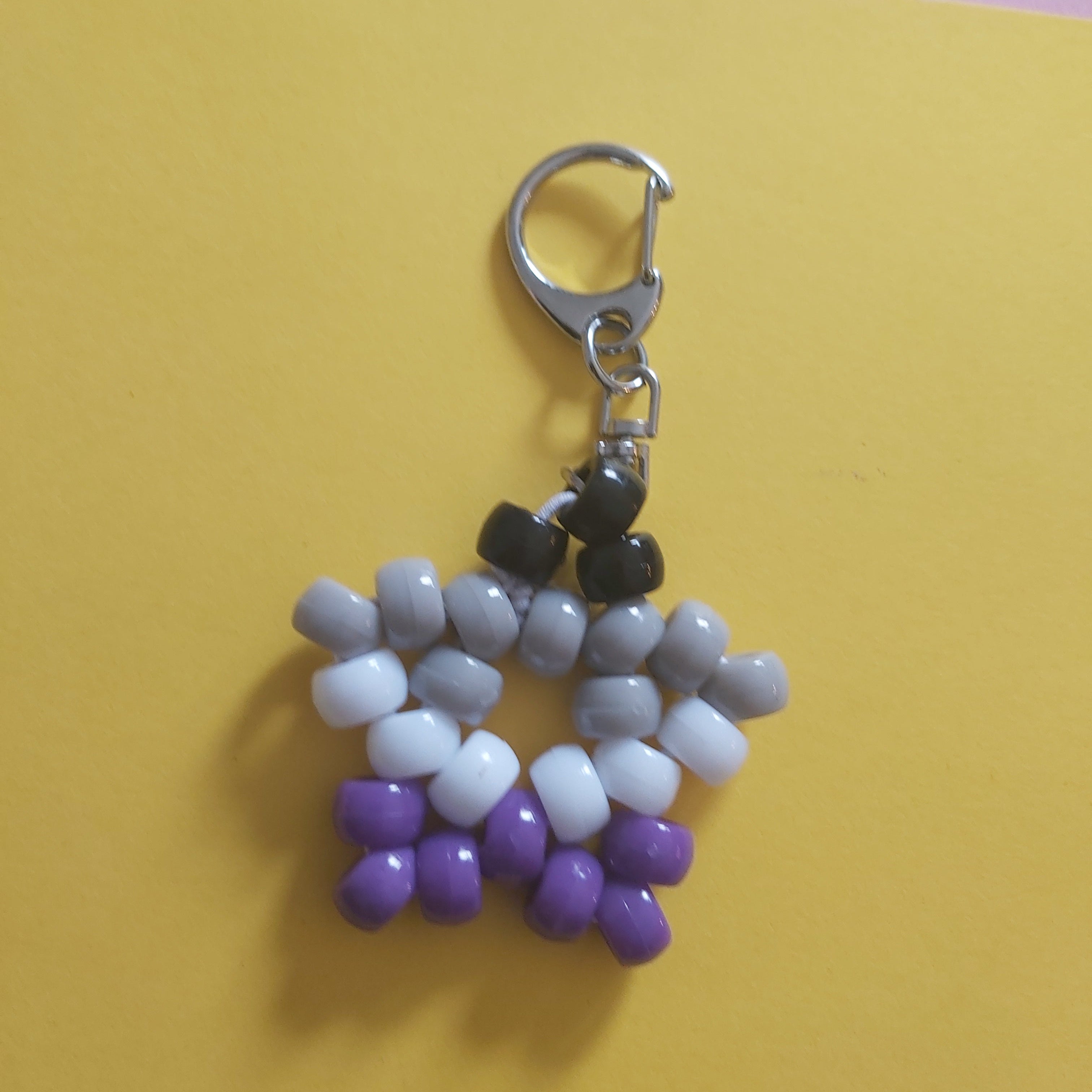 A beaded keychain charm shaped like a star, featuring white and light grey beads on the top half and purple beads on the bottom half, attached to a silver metal lobster clasp, set against a yellow and pink background.