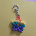 A handmade beaded keychain charm shaped like a rainbow flower, featuring a silver lobster clasp. The beads are arranged in horizontal rainbow stripes of yellow, orange, teal, blue, and purple, resting on a split yellow and purple background.