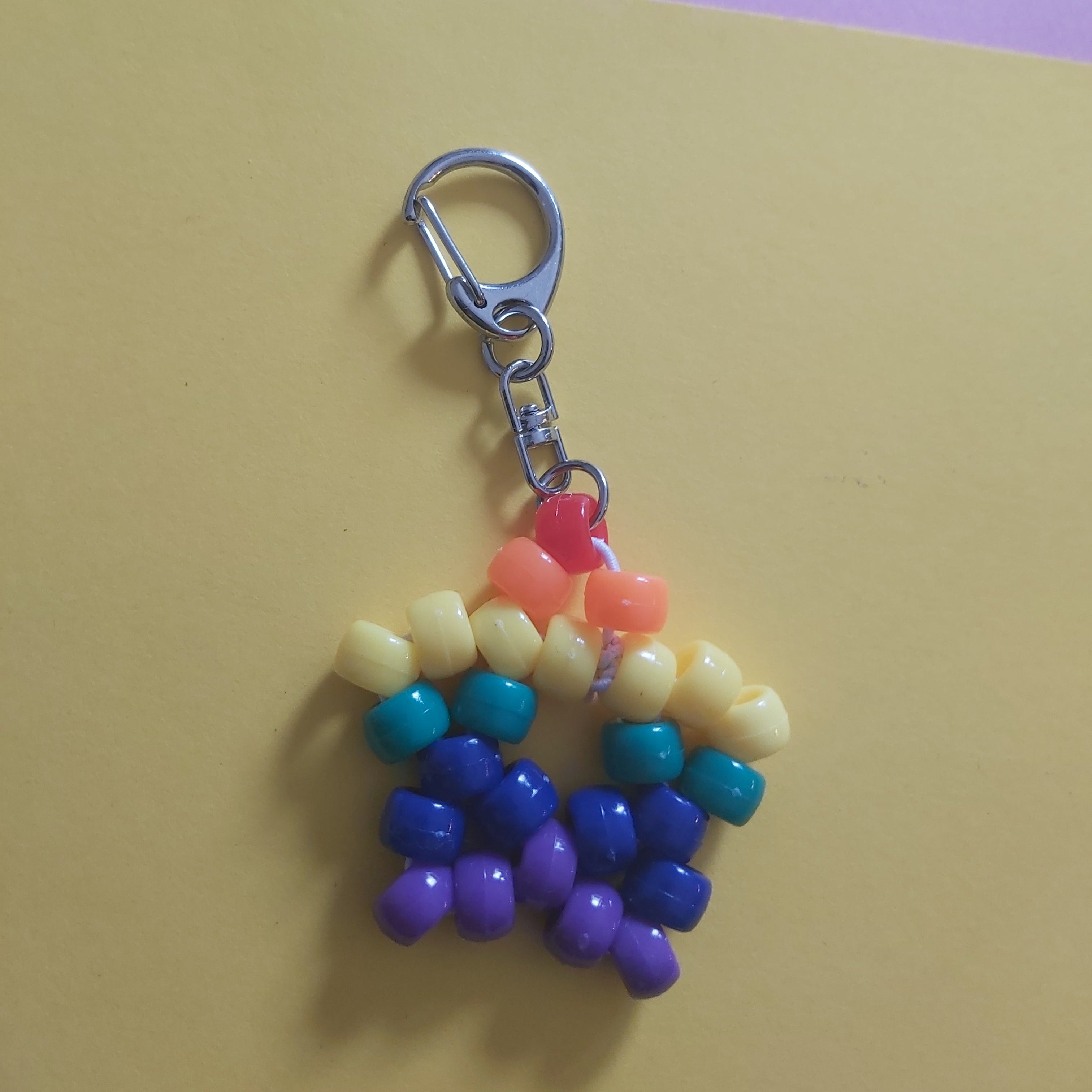 A handmade beaded keychain charm shaped like a rainbow flower, featuring a silver lobster clasp. The beads are arranged in horizontal rainbow stripes of yellow, orange, teal, blue, and purple, resting on a split yellow and purple background.
