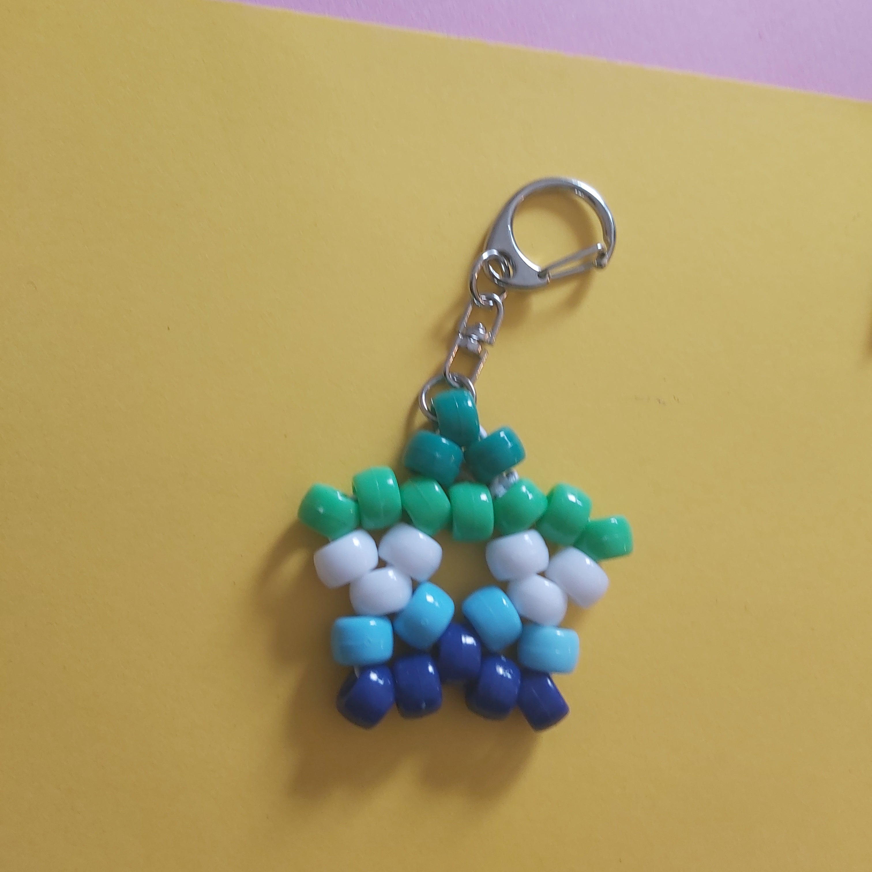 A handmade beaded keychain shaped like a star, made with green, white, light blue, and dark blue beads attached to a silver metal lobster clasp, set against a split yellow and purple background.