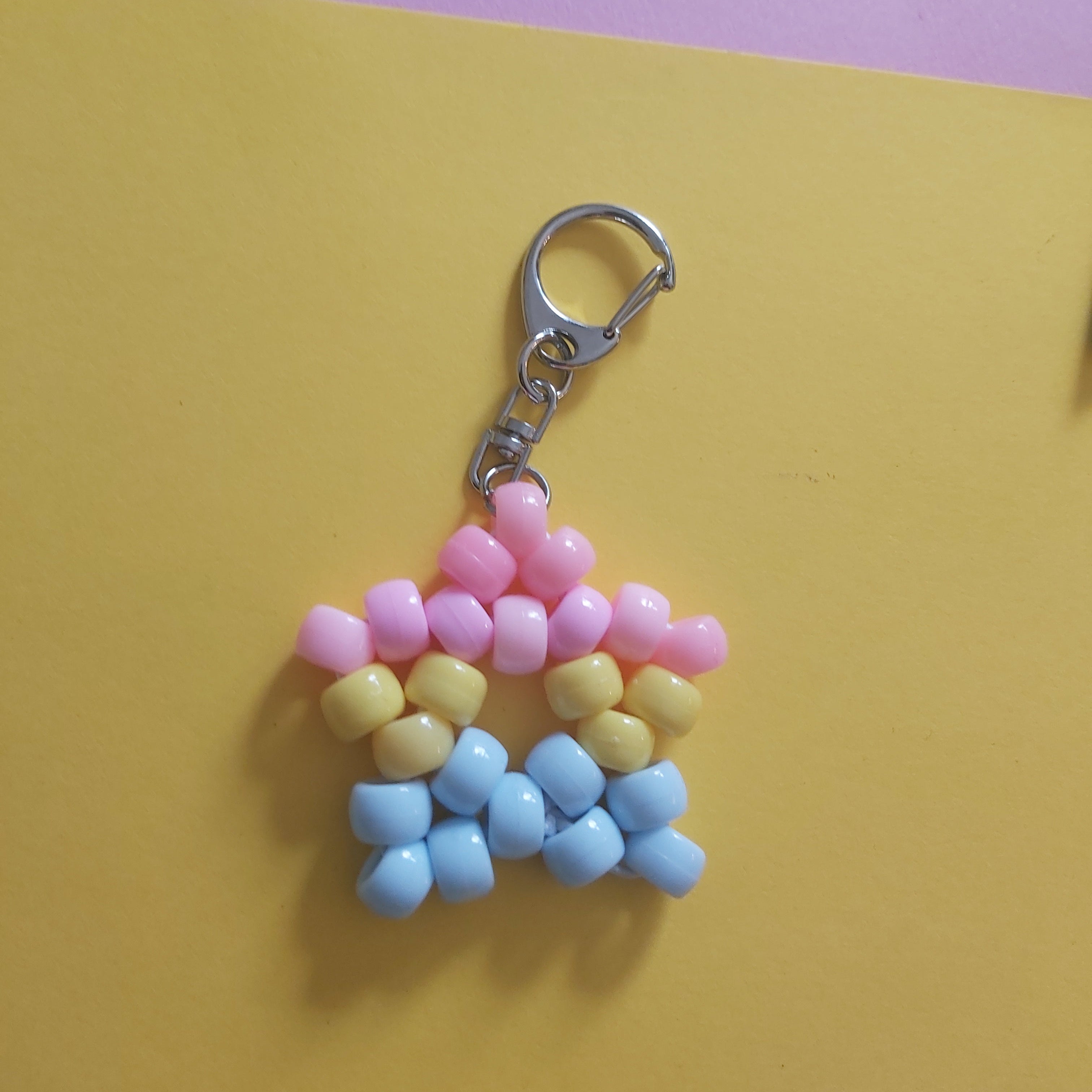 A handmade beaded keychain charm shaped like a star, made with pink, yellow, and light blue beads attached to a silver lobster clasp, set against a split yellow and purple background.