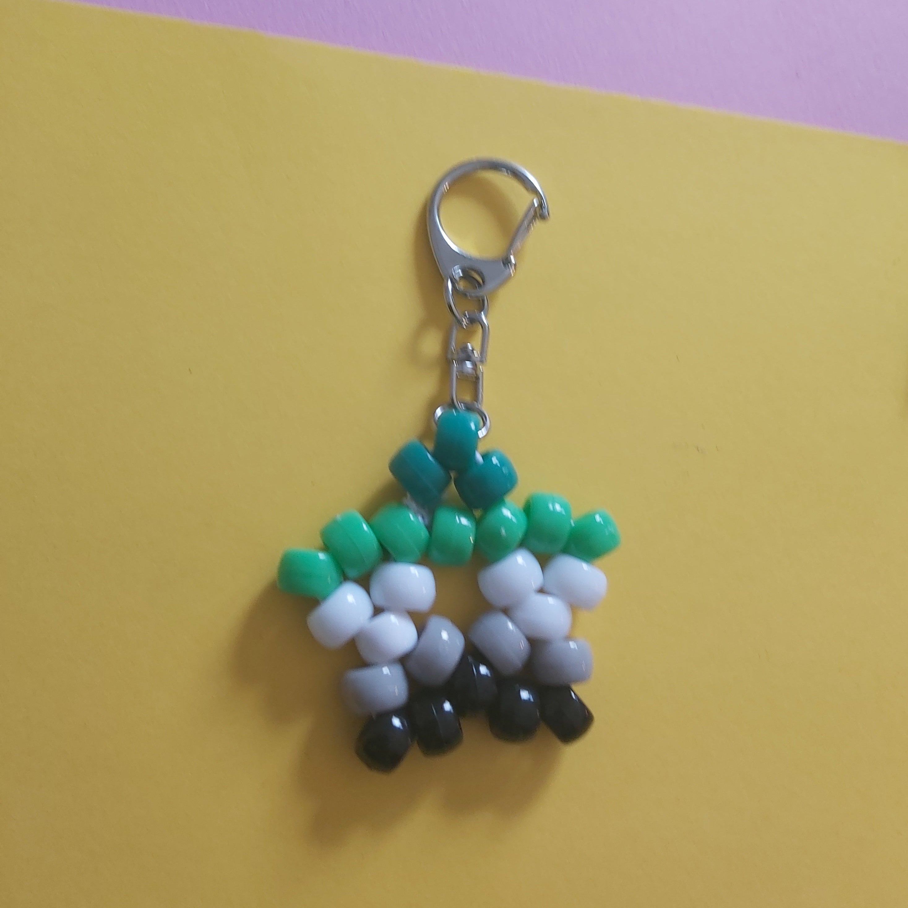A handmade beaded keychain featuring a silver lobster clasp, decorated with green, white, gray, and black beads in a geometric pattern, set against a split yellow and purple background.