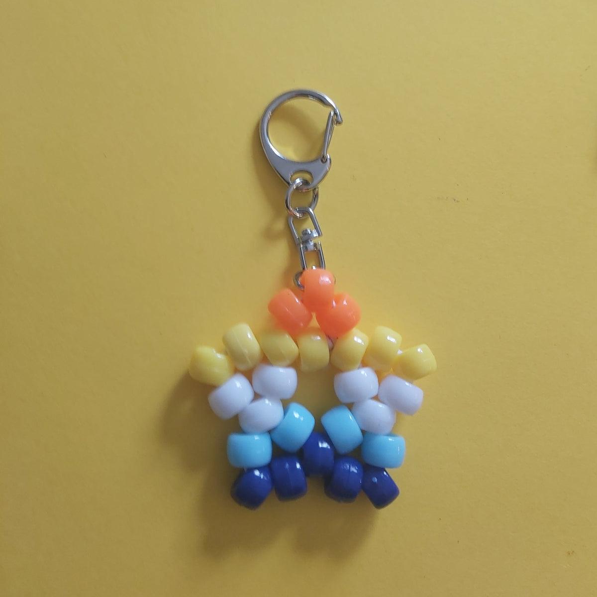A small star-beaded keychain charm with a silver lobster clasp, featuring white, orange, blue, and dark blue beads =, displayed on a yellow and purple background.