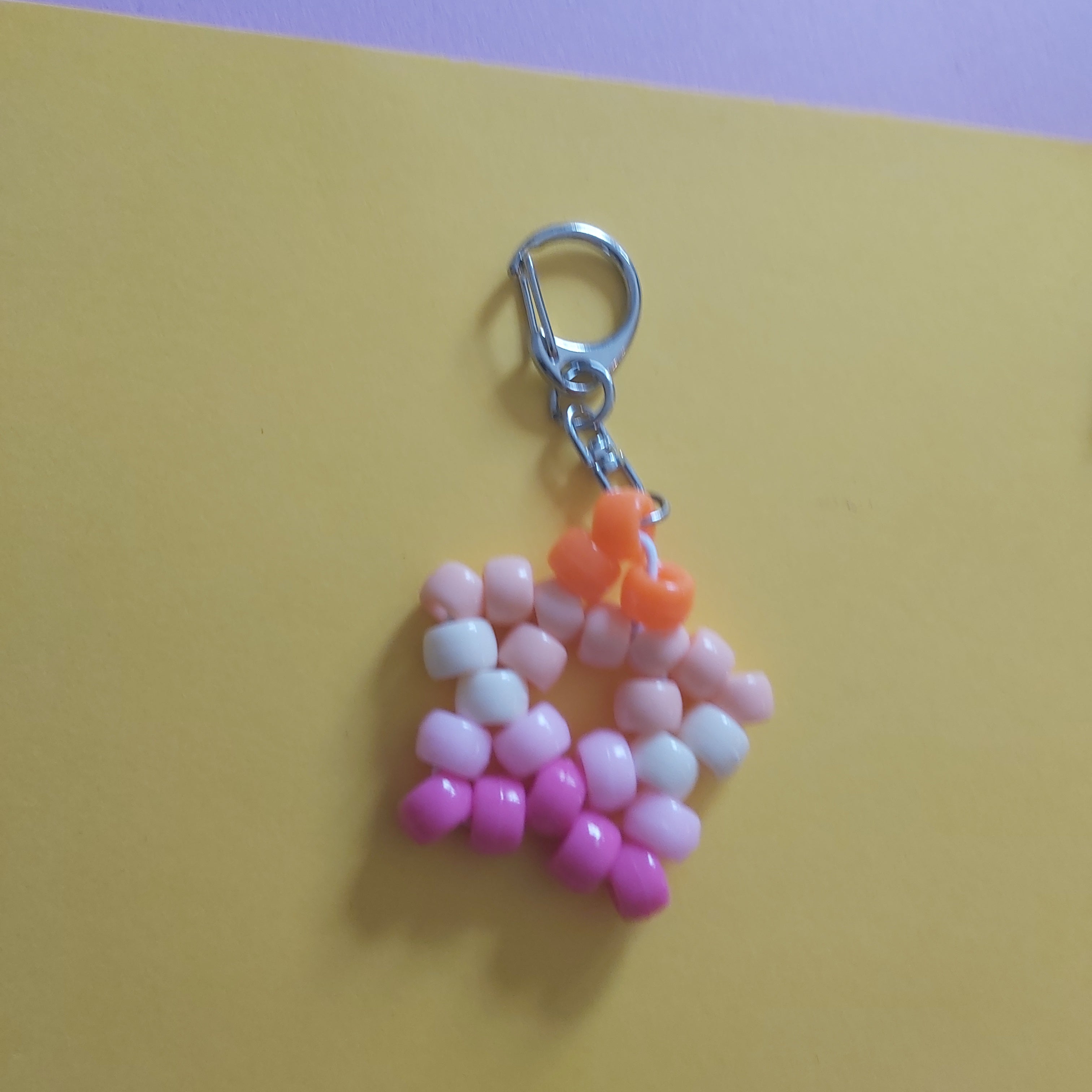 A handmade beaded keychain charm featuring pink, white, and orange beads, attached to a silver lobster clasp, set against a yellow and pink background.