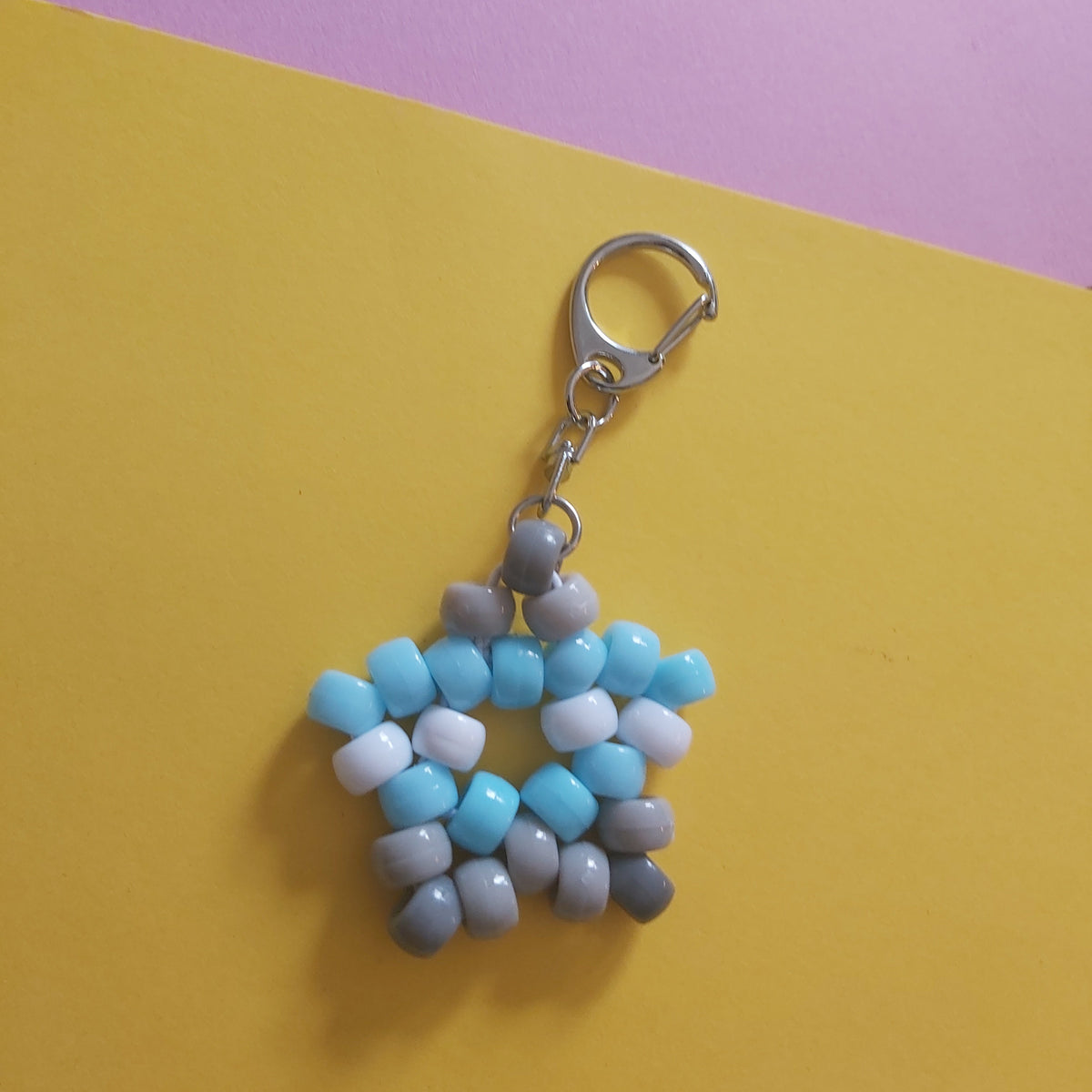 A handmade beaded keychain charm shaped like a star, featuring light blue, white, and grey beads with a silver lobster clasp, set against a split yellow and pink background.