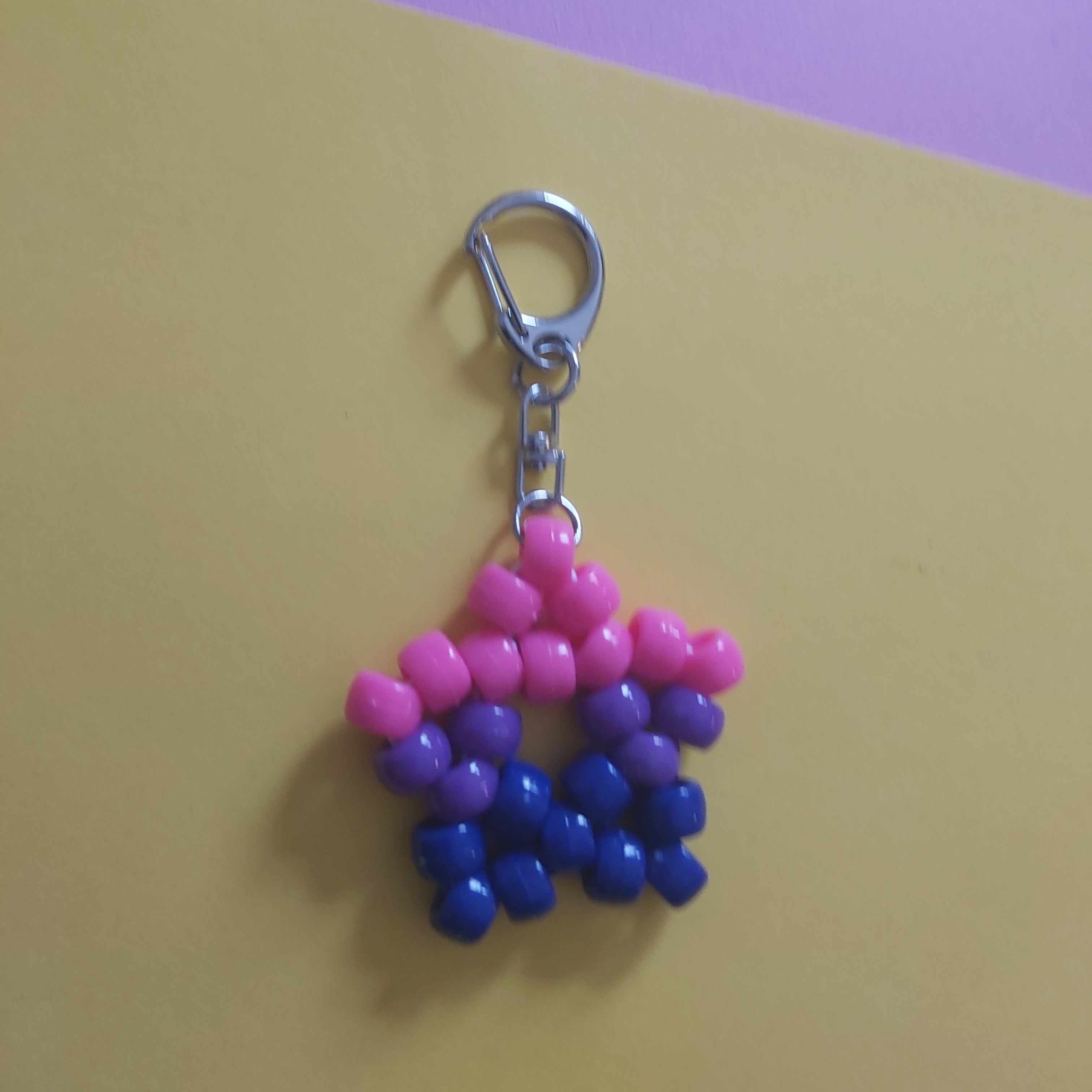 A handmade beaded keychain charm in the pink, blue and purple stripes of the bisexual pride flag, attached to a silver metal lobster clasp, displayed on a yellow and purple background.