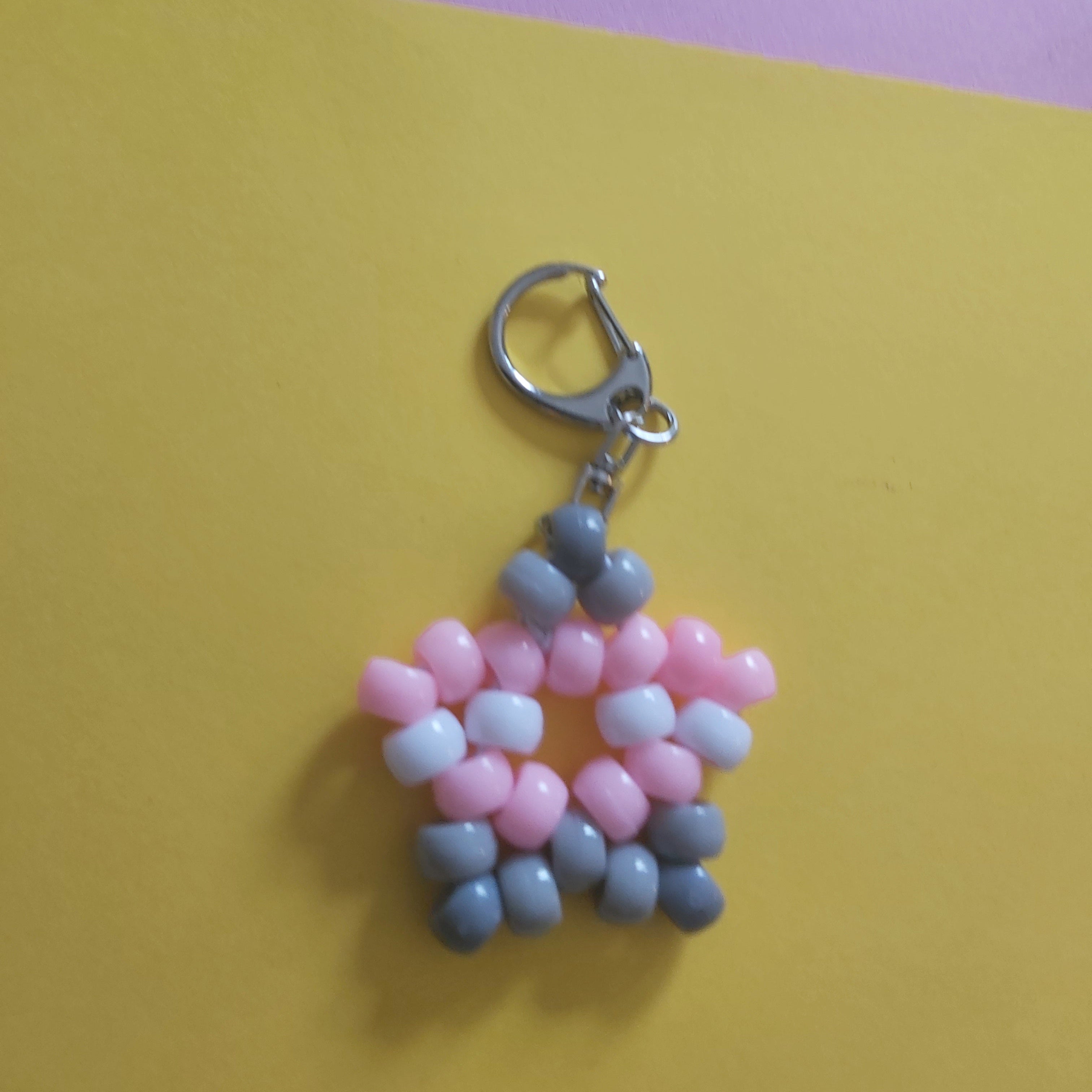 A handmade beaded keychain charm shaped like a star with pink, white, and grey beads, attached to a silver metal lobster clasp, set against a split yellow and purple background.