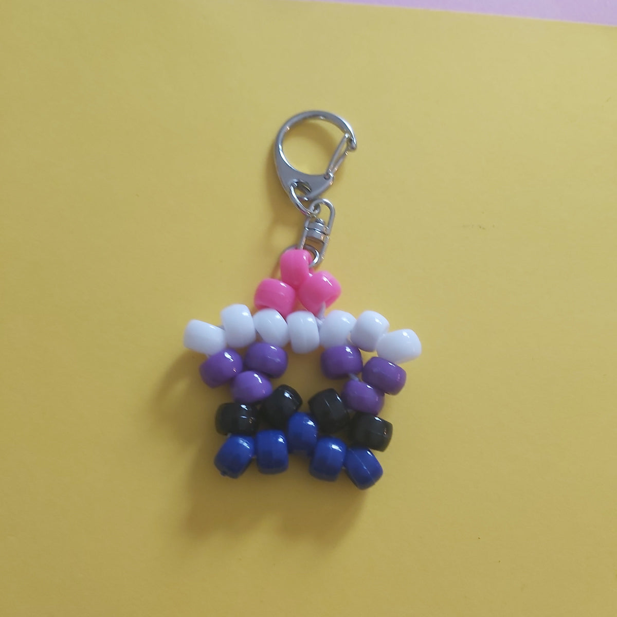 A handmade beaded keychain charm shaped like a star, featuring pink, white, purple, black, and blue beads with a silver metal clasp, set against a yellow and pink background.