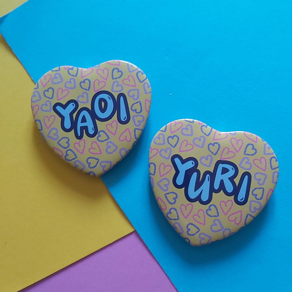 Two heart-shaped pins with colourful heart patterns and the names 'YAOI' and 'YURI' in blue bubble letters, displayed on a background of yellow, pink, and blue paper.