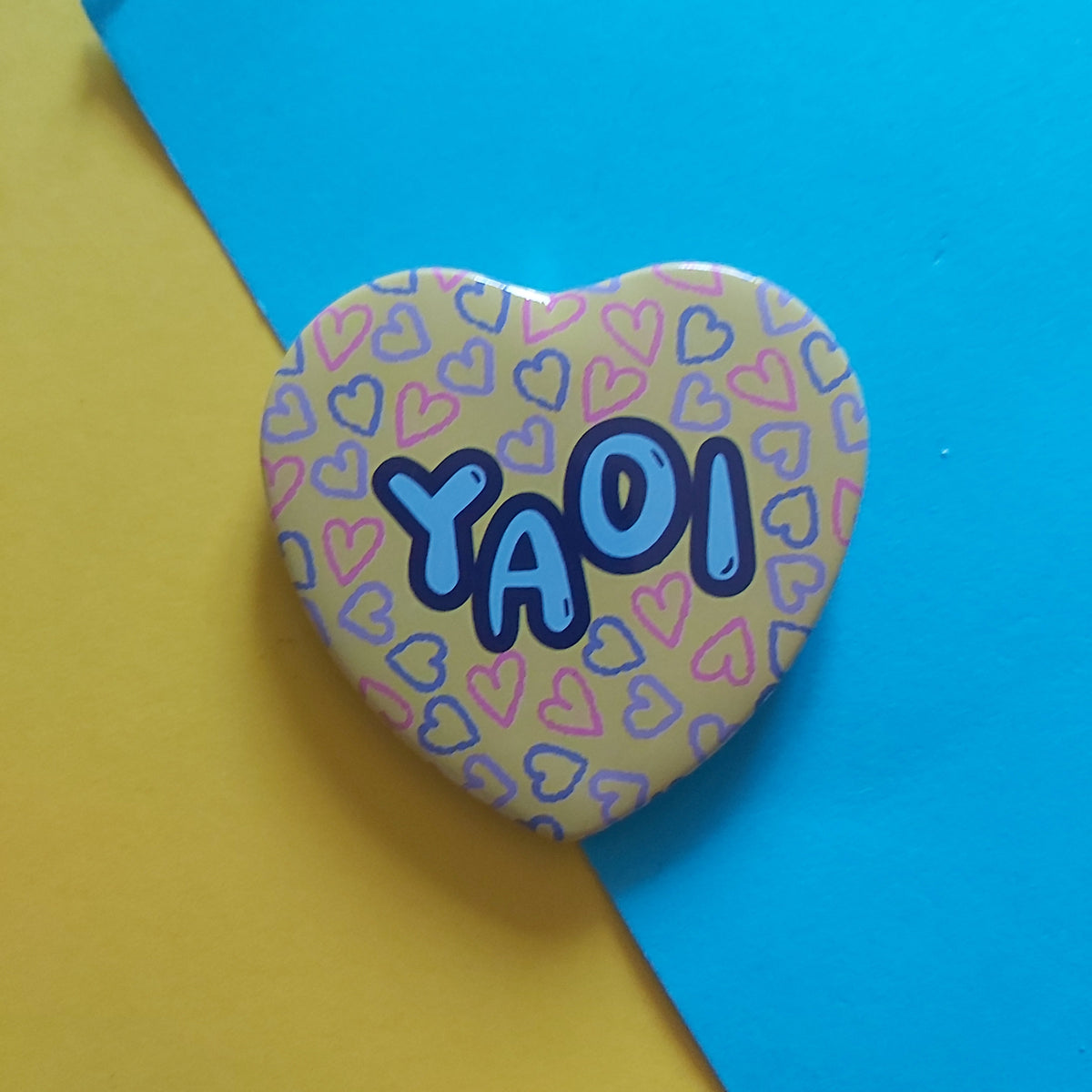 A heart-shaped pinback button featuring the word 'YAOI' in blue bubble letters over a pattern of small hearts, displayed on a diagonal split yellow and blue background.
