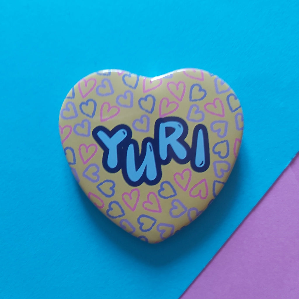 A heart-shaped pinback button badge featuring the name 'YURI' in blue bubble letters with a dark blue outline, set against a pale yellow background patterned with small pink and blue hearts. The badge is photographed on a vibrant blue background with a pink corner accent.