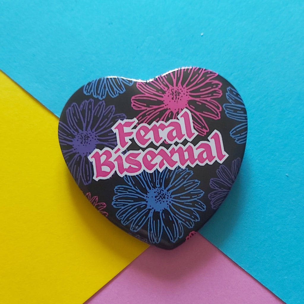 A heart-shaped pin with a black background, pink and blue floral pattern, and the text 'Feral Bisexual' in pink, displayed on a colourful background of yellow, blue, and pink paper.