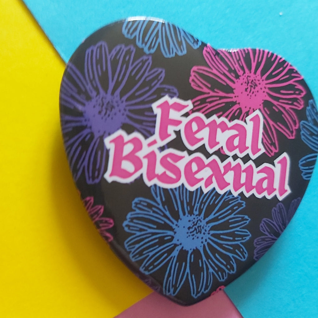 A heart-shaped pinback button featuring the text 'Feral Bisexual' in pink with a white outline, set against a black background with pink, purple and blue daisy illustrations. The button is displayed on a colourful background of yellow, blue, and pink paper.