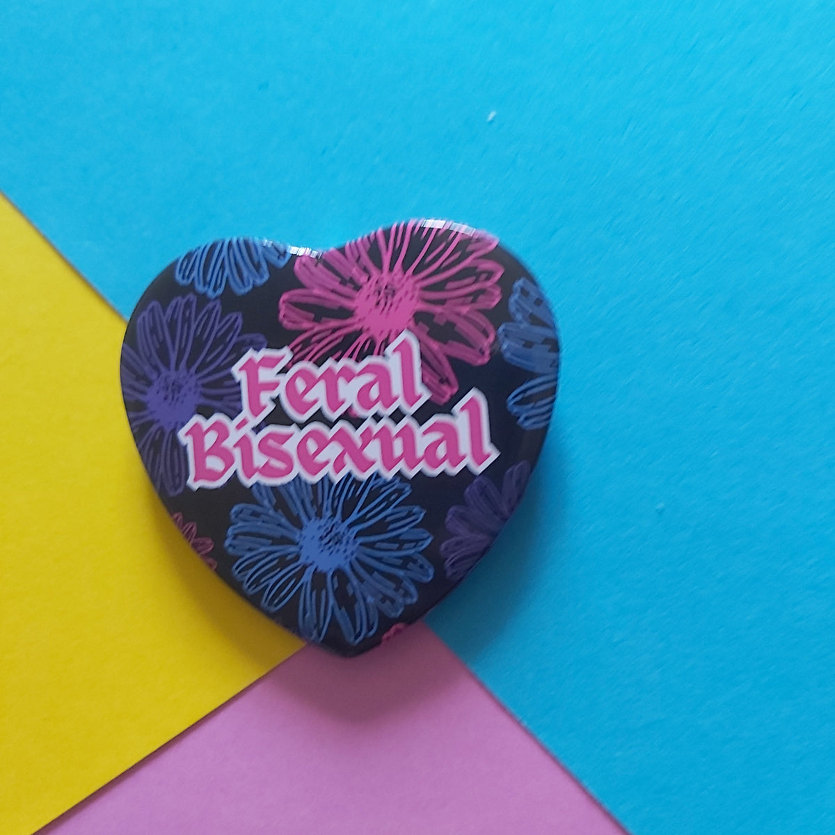 A heart-shaped pin with a floral pattern and the text 'Feral Bisexual' in pink, placed on a background divided into yellow, blue, and pink sections.