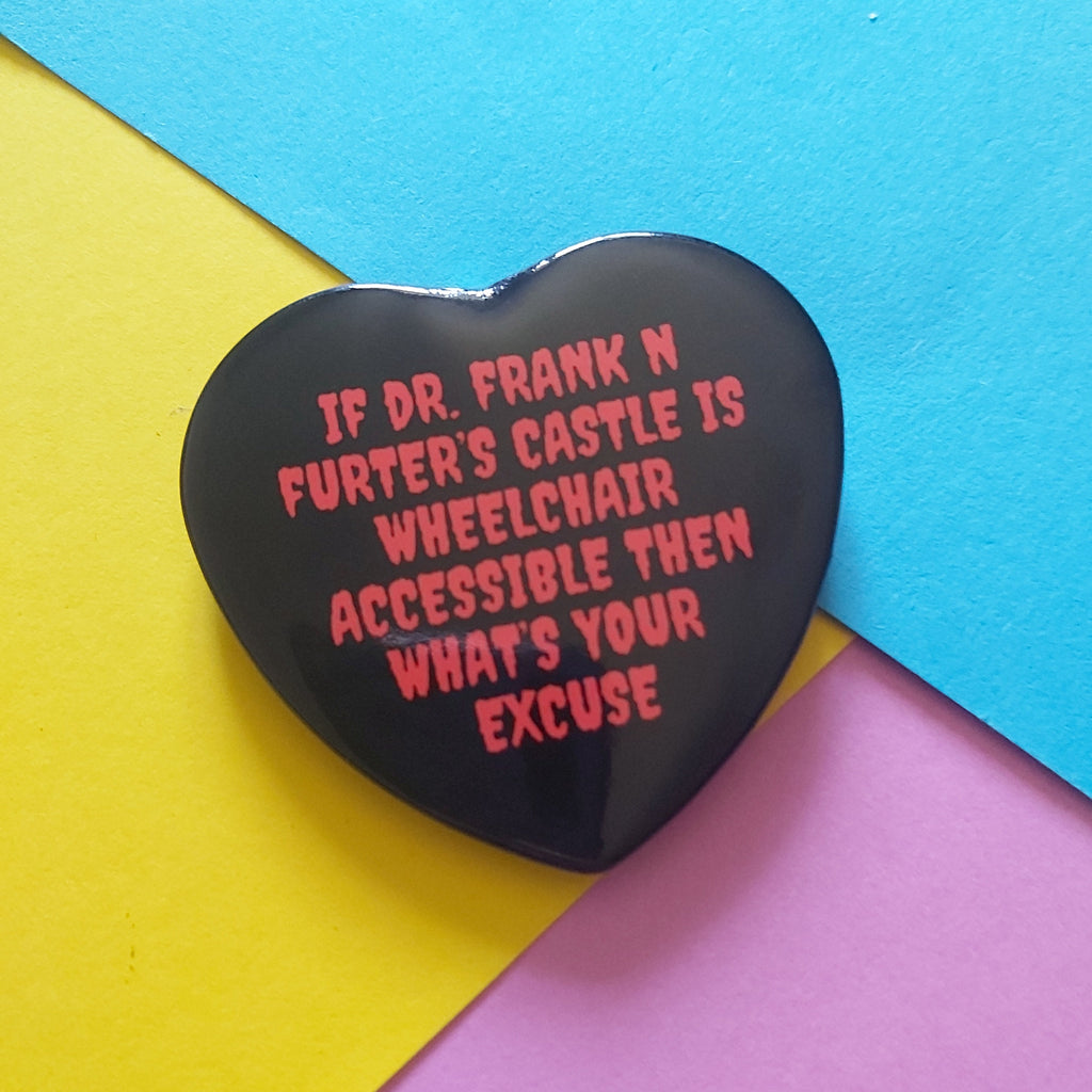 A heart-shaped black pin with red text reading 'IF DR. FRANK N FURTER'S CASTLE IS WHEELCHAIR ACCESSIBLE THEN WHAT'S YOUR EXCUSE', photographed on a colourful background of yellow, blue, and pink paper.